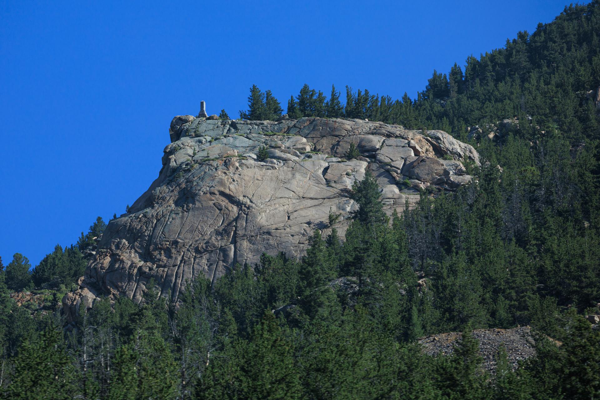 Rocky outcrop with a white marker on top, surrounded by dense green forest.