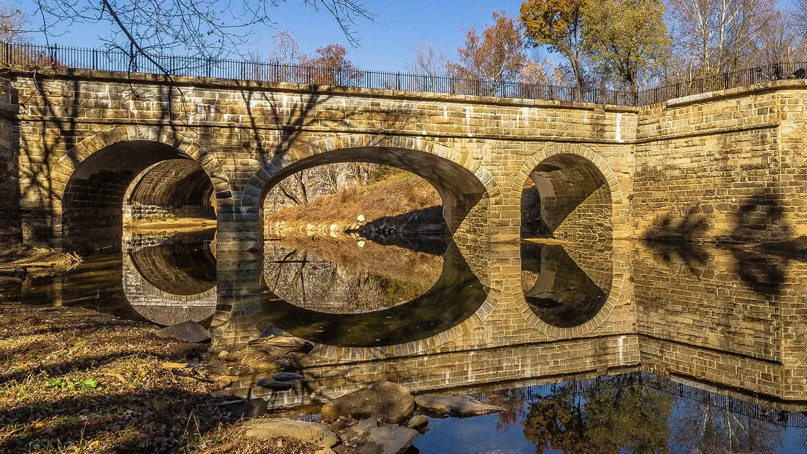An old stone bridge with arches reflects in the water on a sunny day.