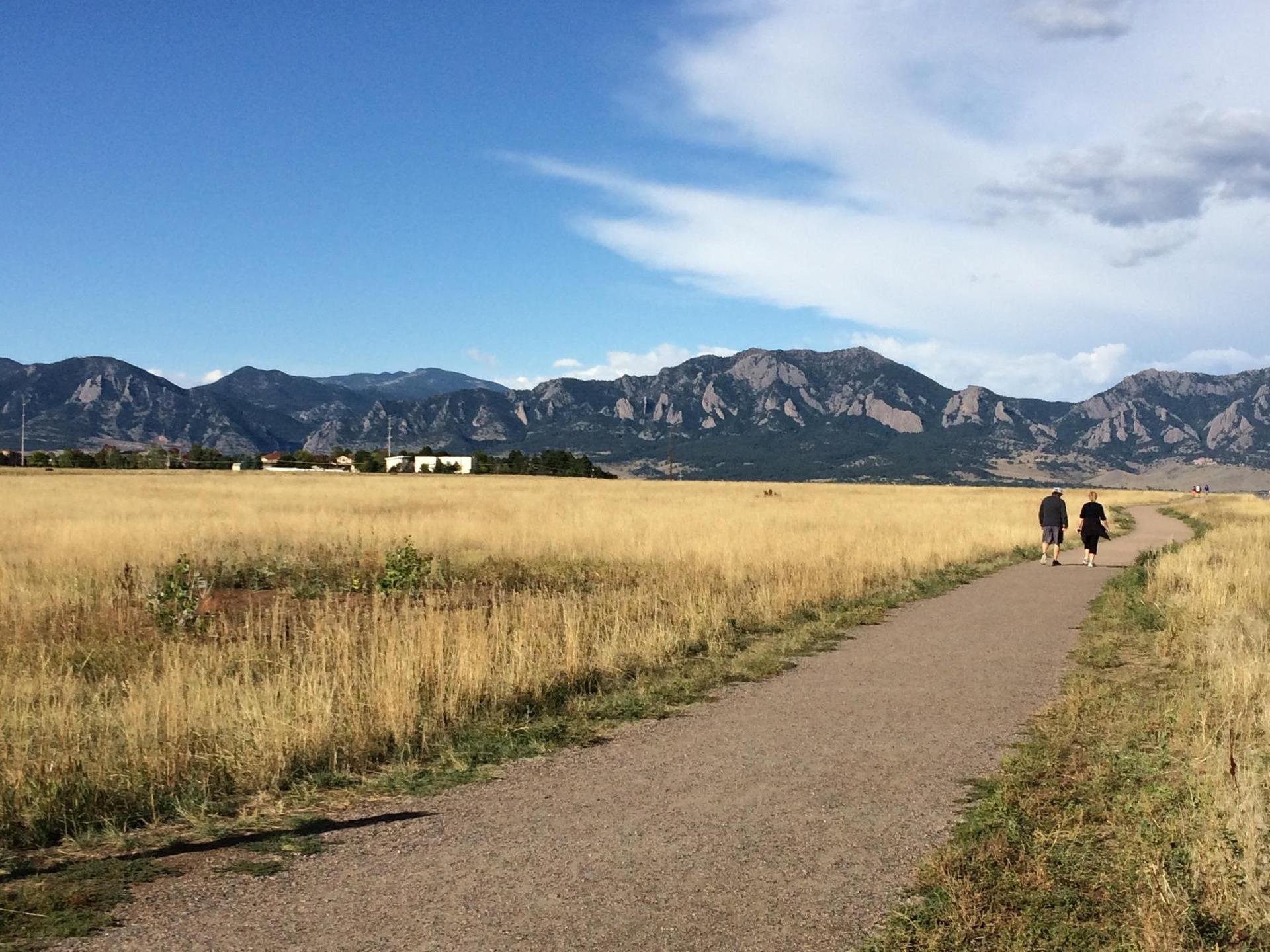 Two people walk on a path through golden fields with mountains in background.