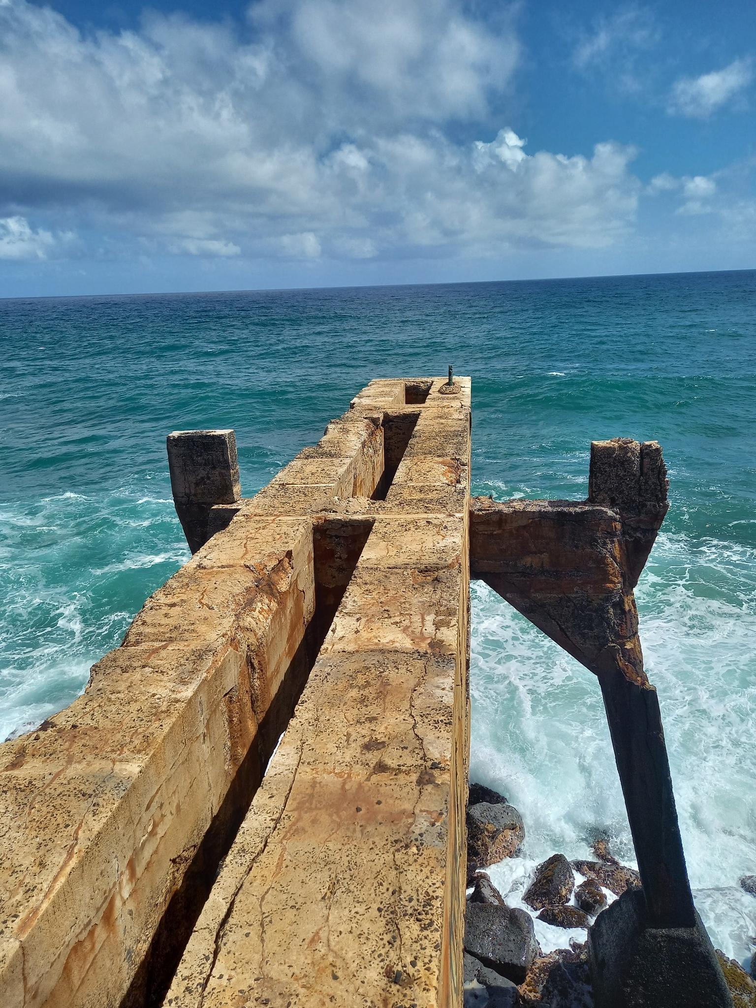 An old concrete pier extends into the blue ocean with crashing waves.