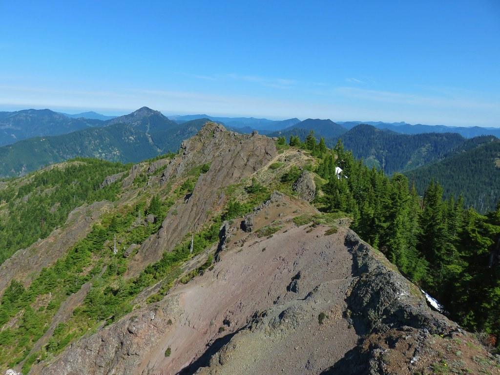 A rocky mountain ridge under a clear blue sky, surrounded by green peaks.