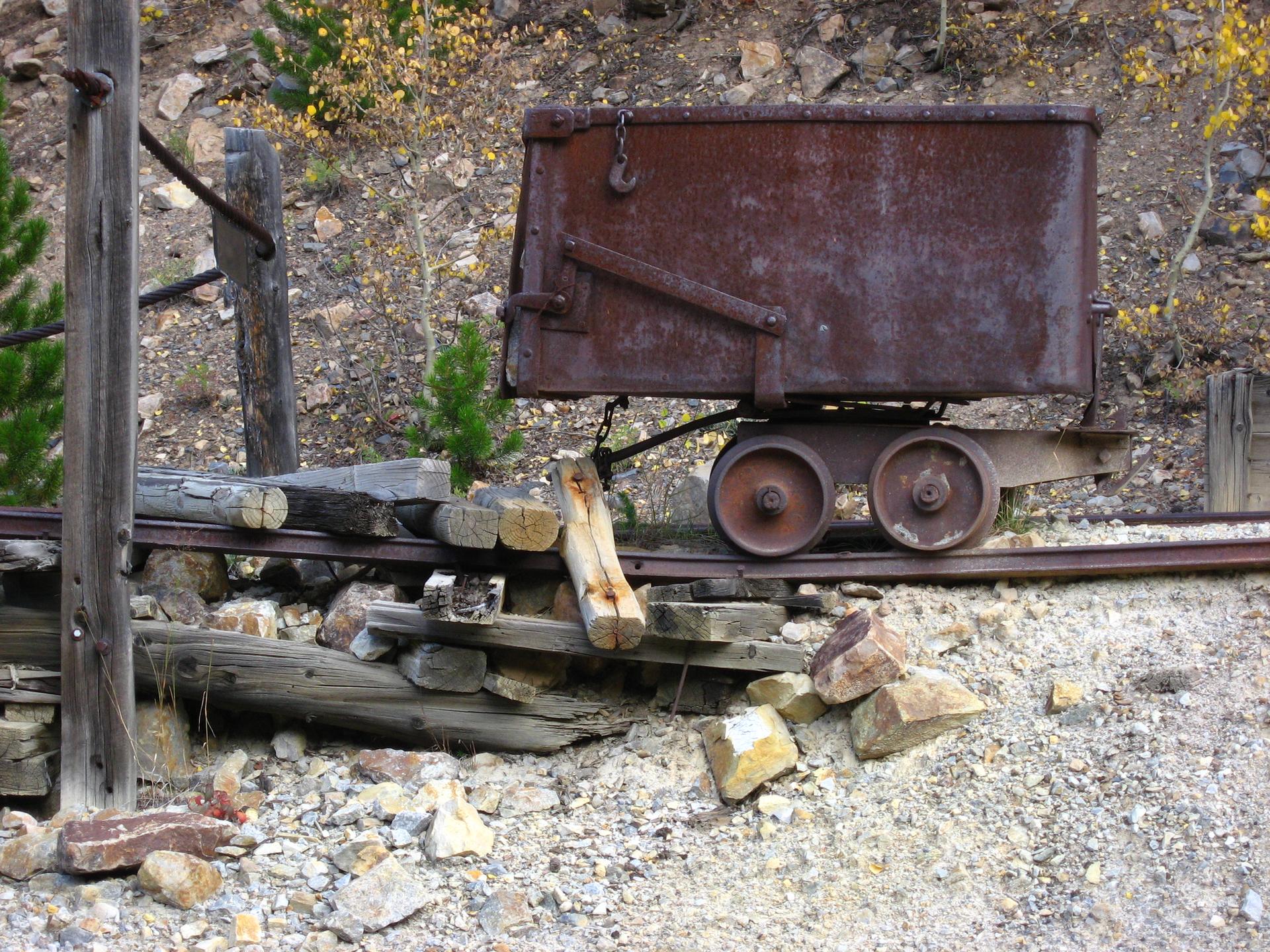 Old rusty mining cart on tracks amidst rocks and sparse trees in daylight.