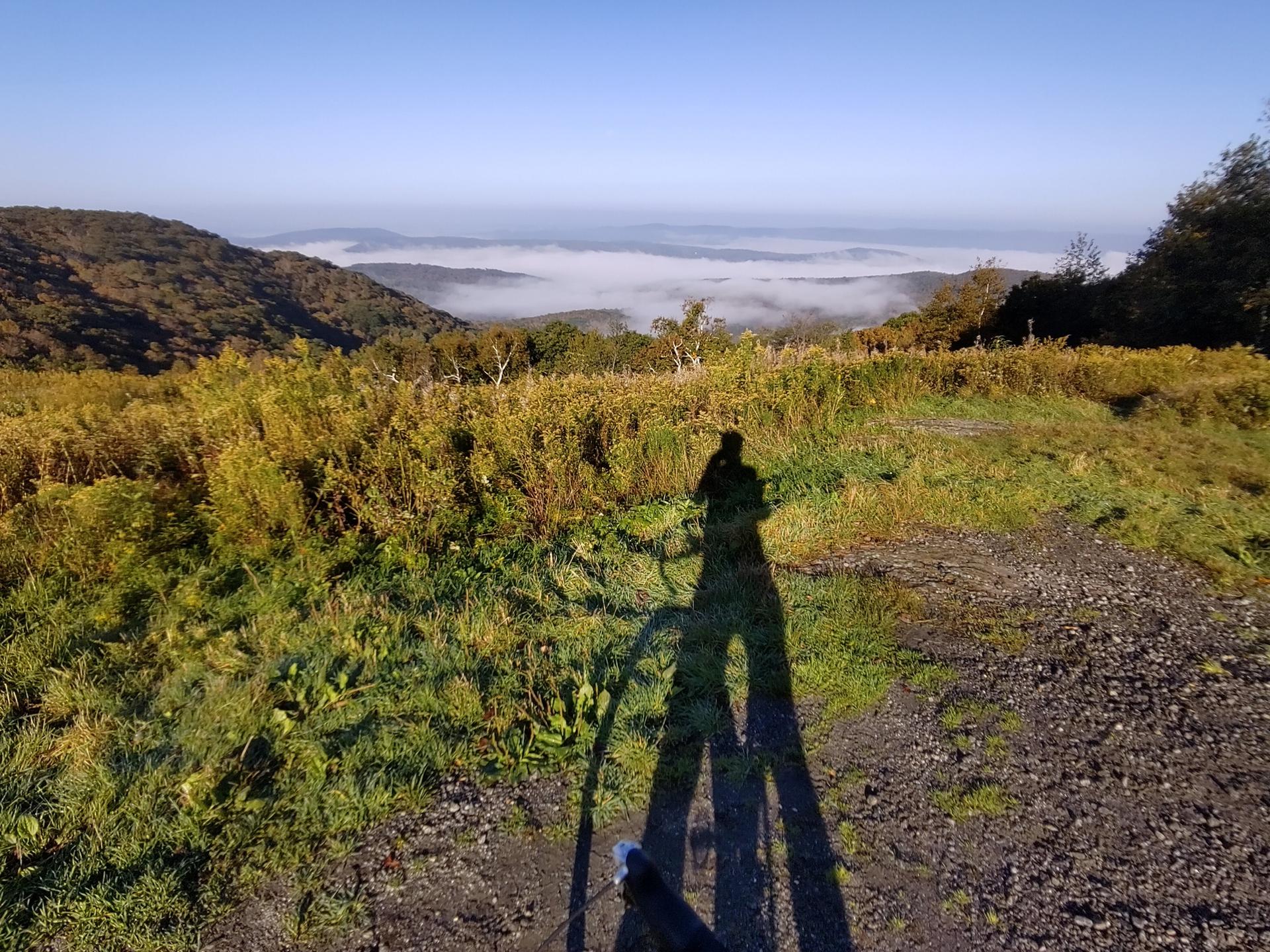 Long shadow of person and bike on a grassy hill overlooking foggy valley.