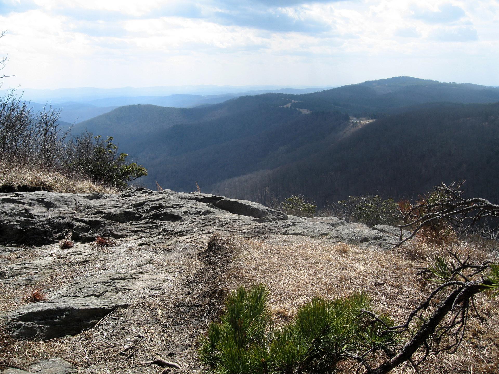 Panoramic mountain vista from a rocky cliff under a partly cloudy sky.