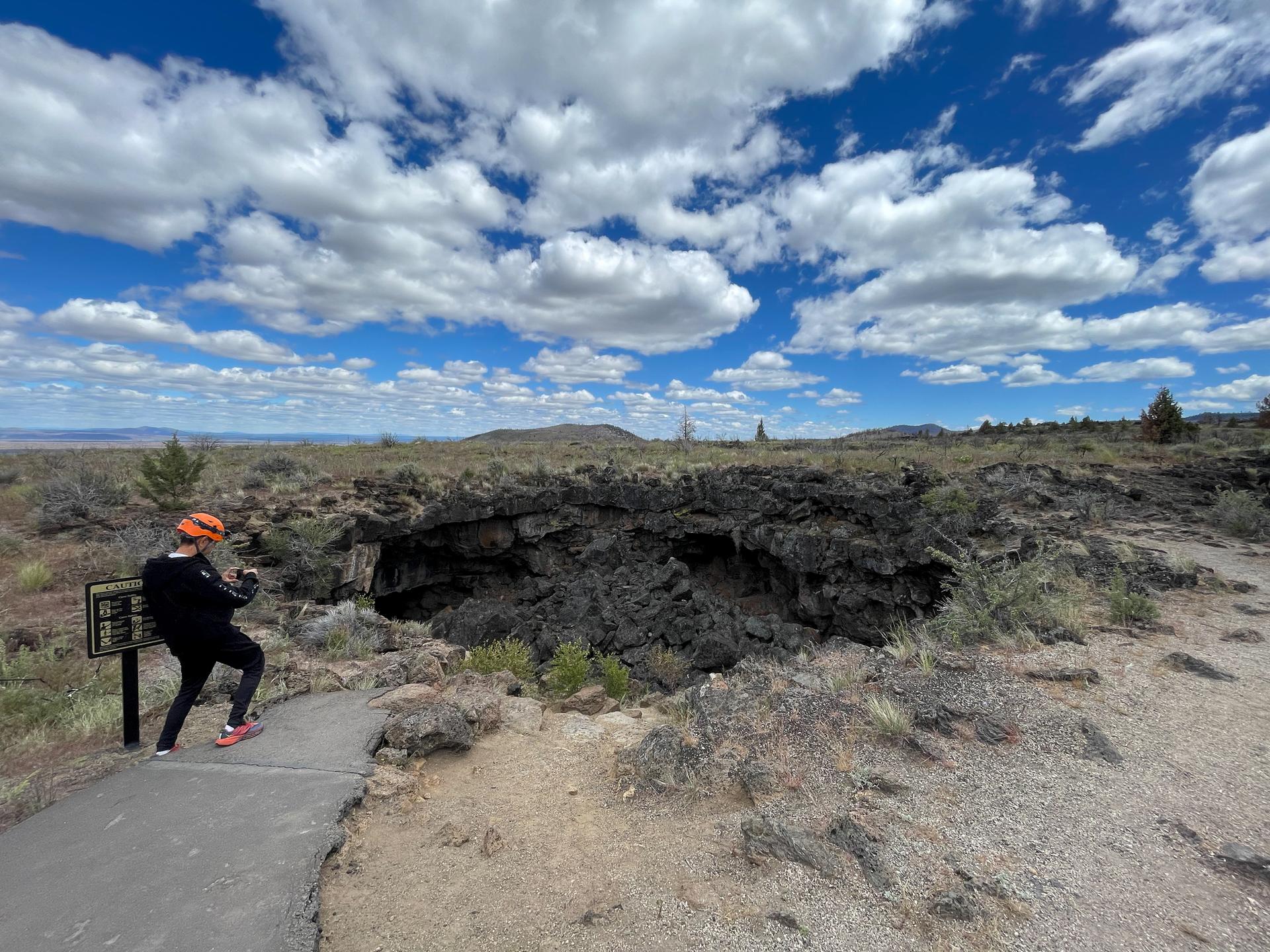 Person in helmet near a dark cave entrance under a cloudy sky.