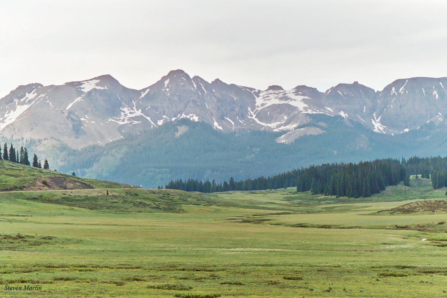 Wide green meadow with forested hills and distant snow-capped mountains under an overcast sky.