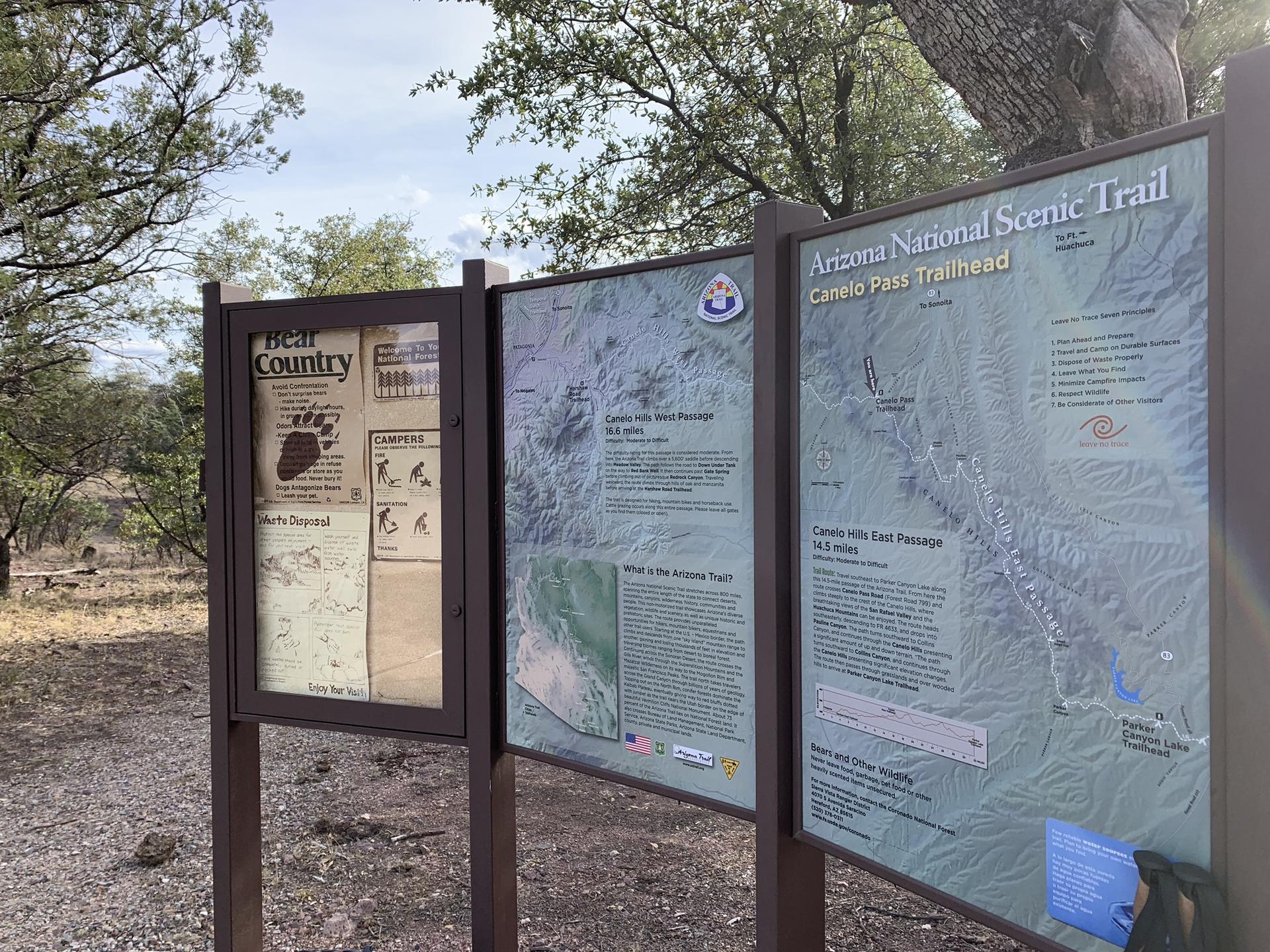 Three information kiosks with maps and text at a trailhead.