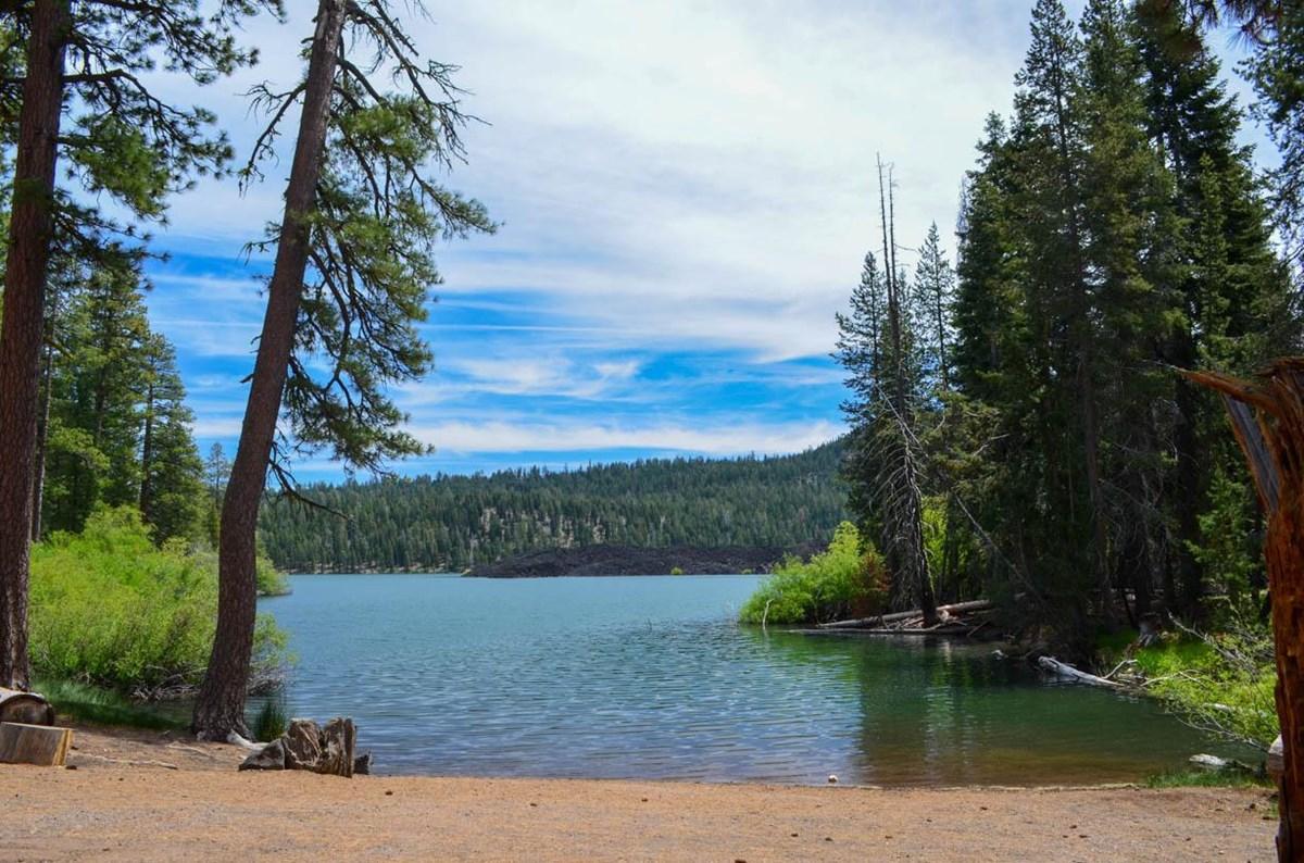 A tranquil lake surrounded by a dense forest under a bright, partly cloudy sky.