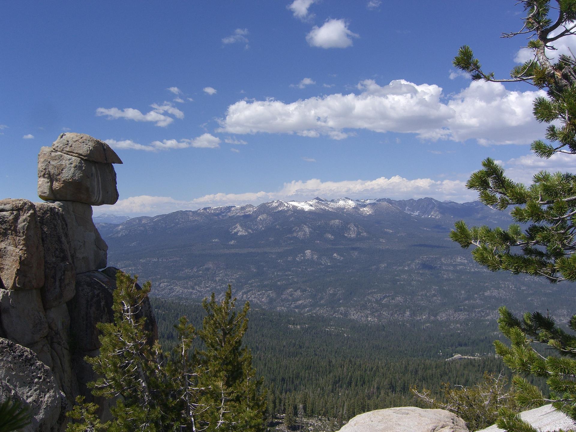 A balanced rock formation overlooks a vast, sunny, snow-capped mountain range.