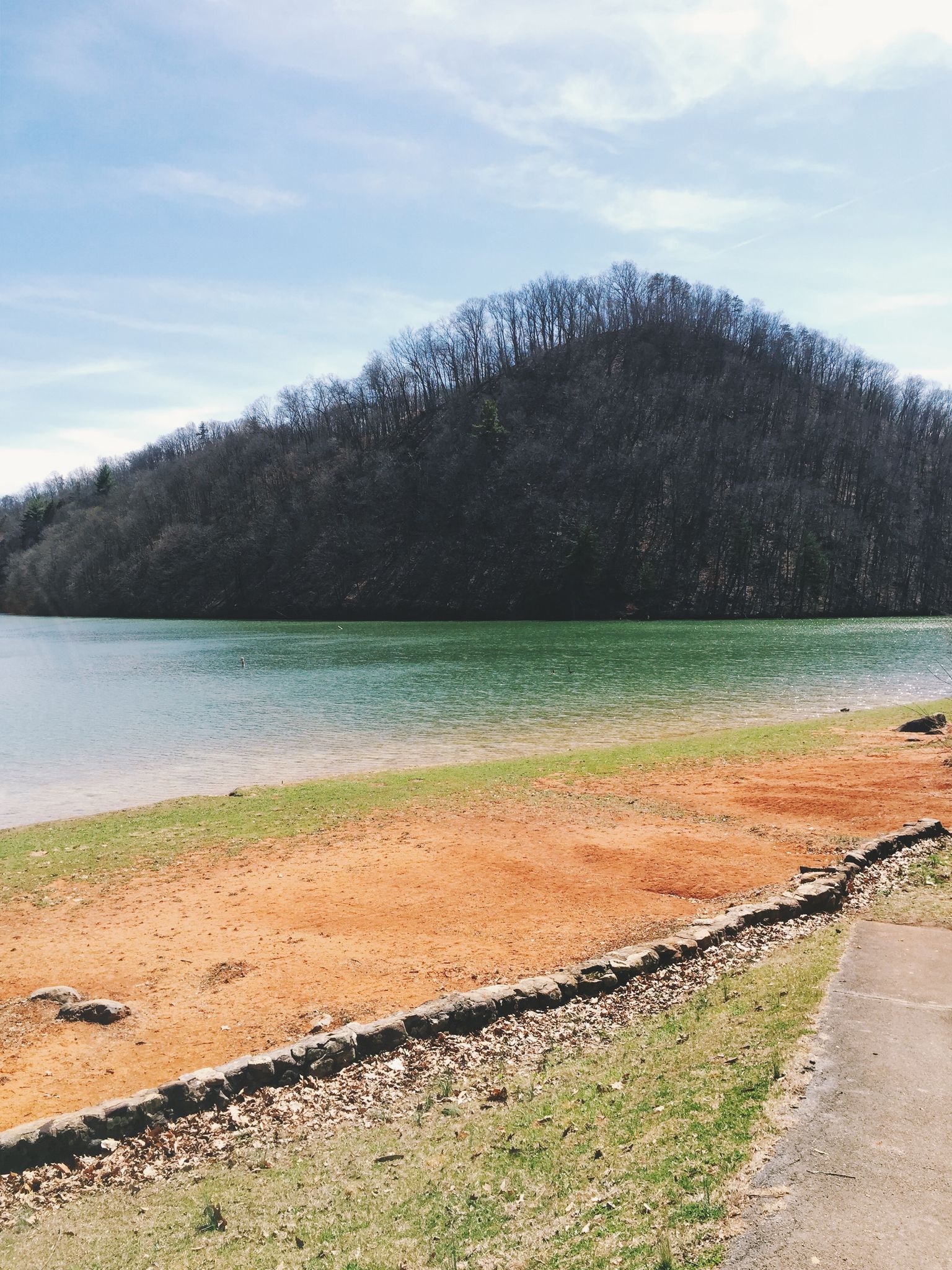 A calm lake with a reddish-brown shore and a forested hill under a bright sky. - Lakeside Trail loop from McDonald's