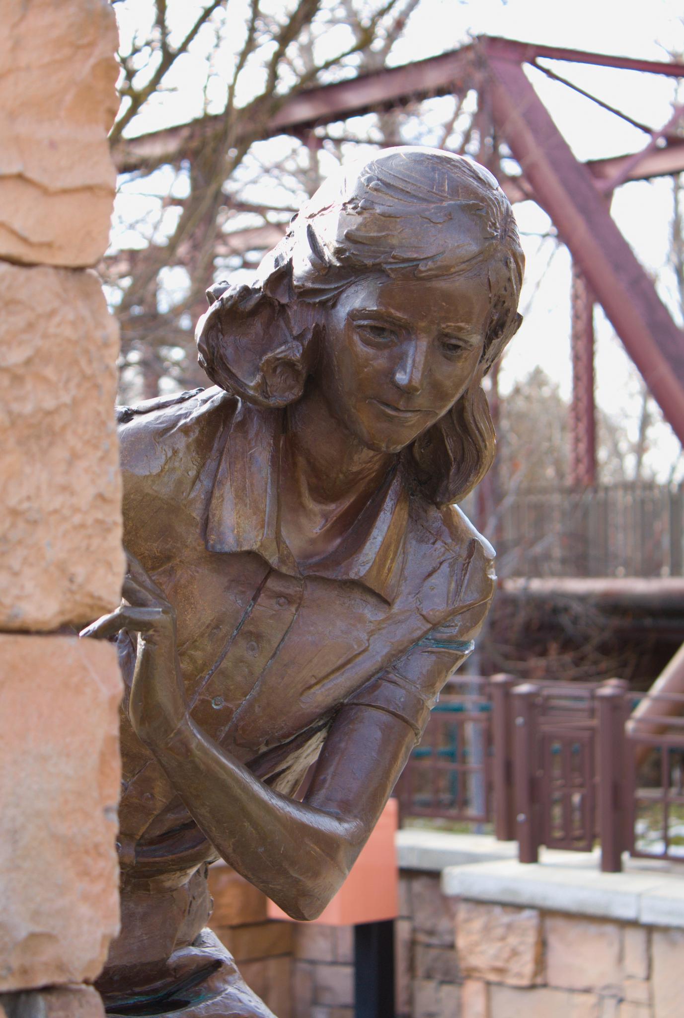 Bronze statue of a young woman peeking from behind a stone wall.