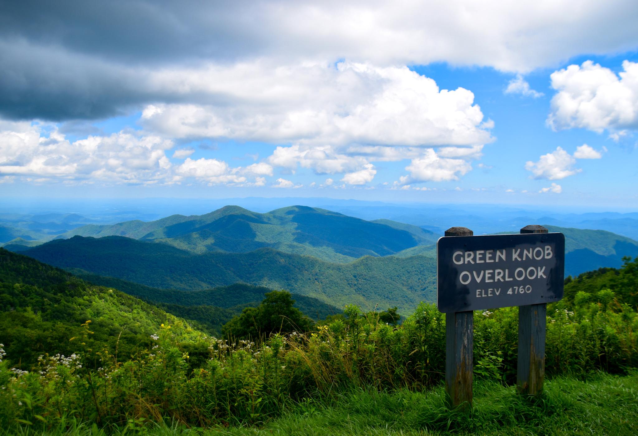 A wooden overlook sign in foreground with lush green mountains under a cloudy sky.