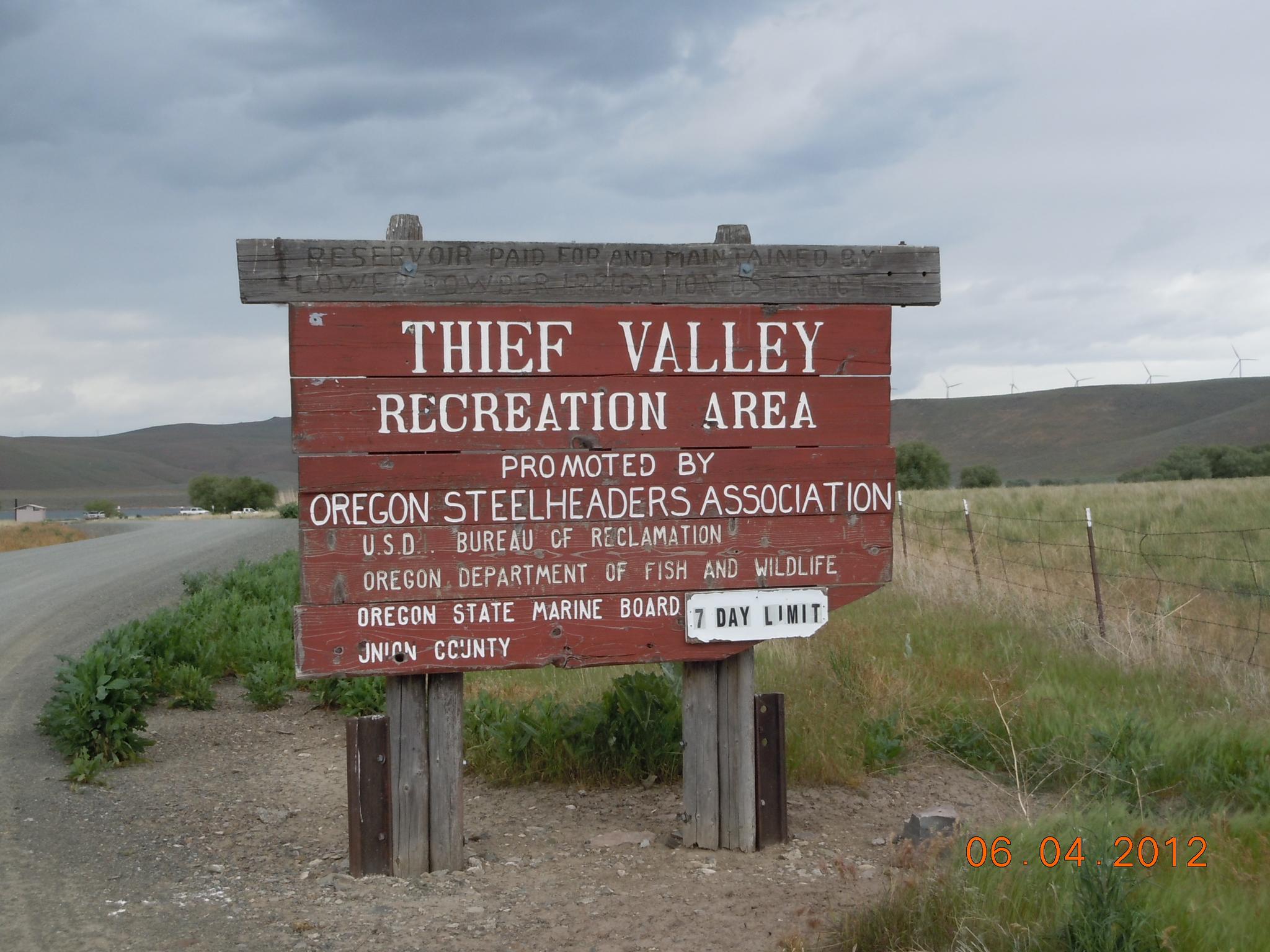 A rustic wooden sign stands beside a dirt road in a rural landscape.