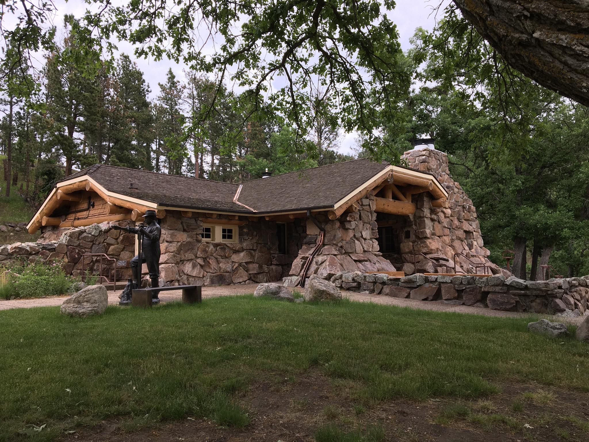 Rustic stone and log building with a statue by a water pump. - Grace Coolidge Creekside Trail