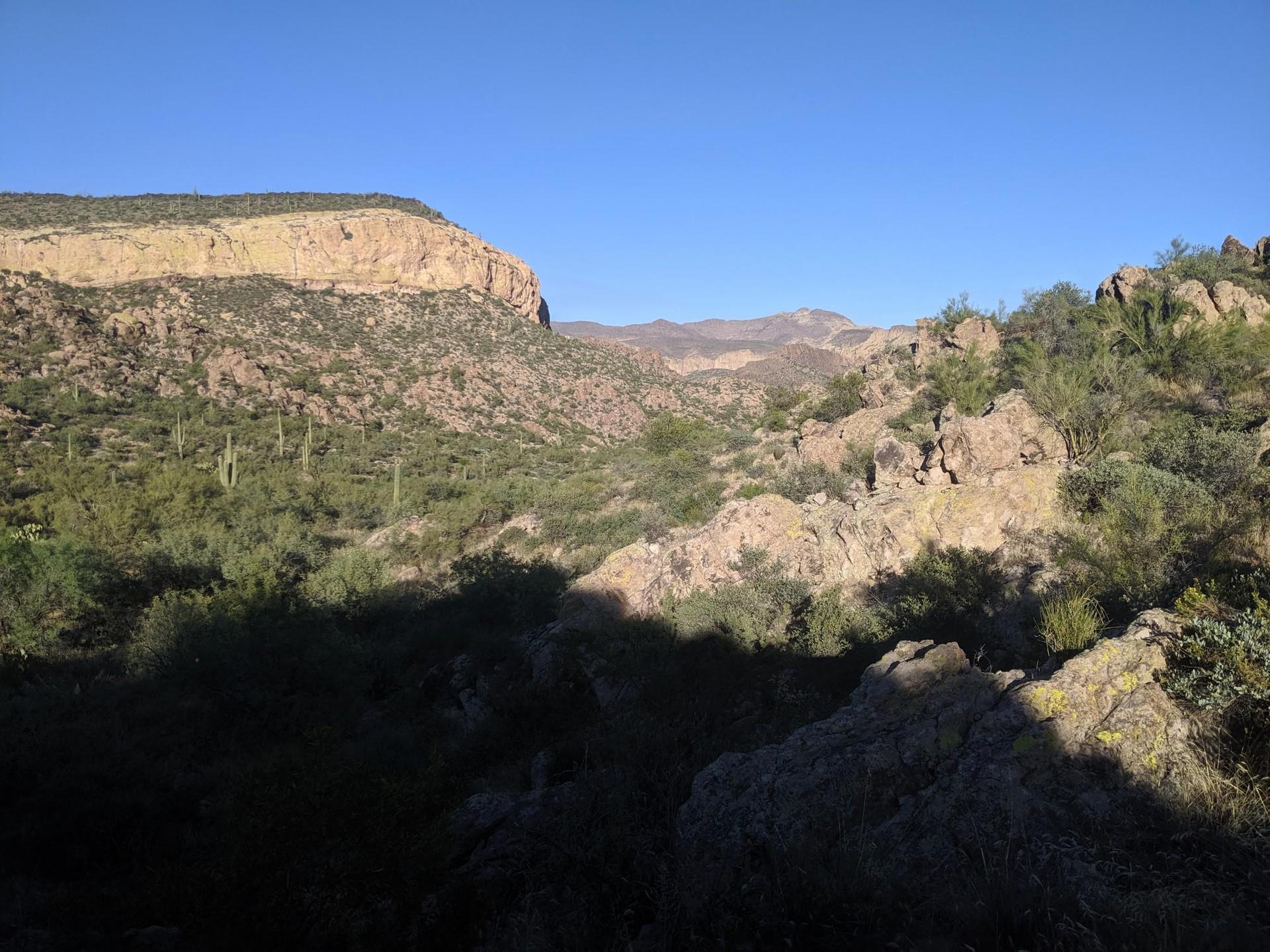Rocky desert valley with green scrub and distant mountains, bright sky.
