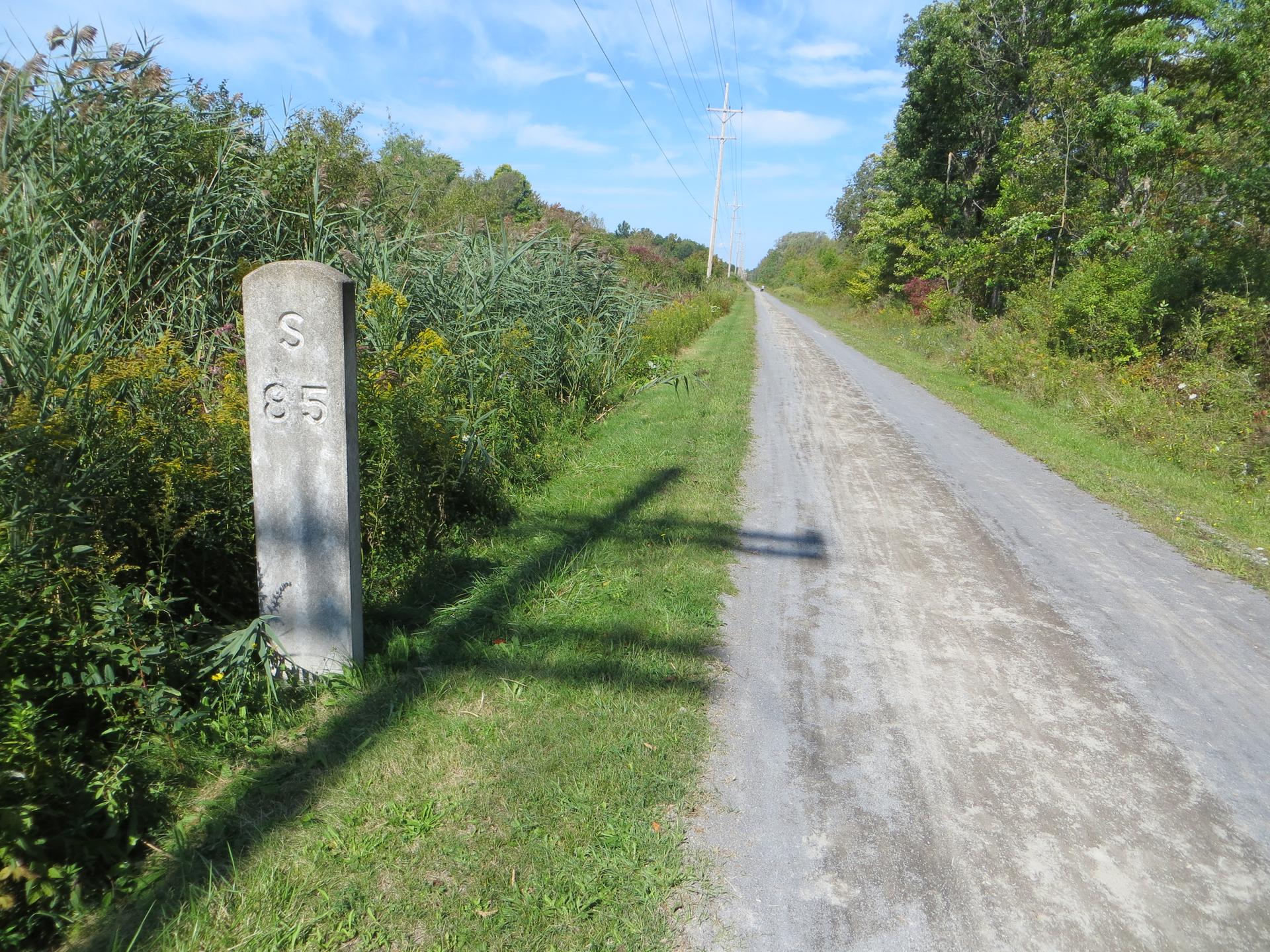 Concrete marker post next to a gravel path through tall grass on a sunny day.