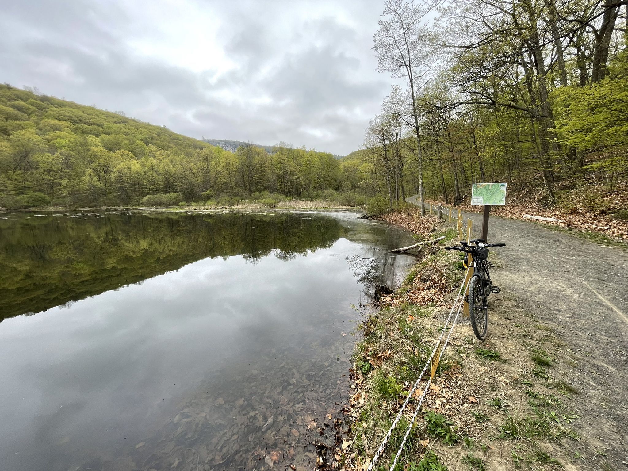 Calm lake beside a dirt path, surrounded by green forest, with a bicycle. - Duck Pond – Duck Pond loop from Town of New Paltz
