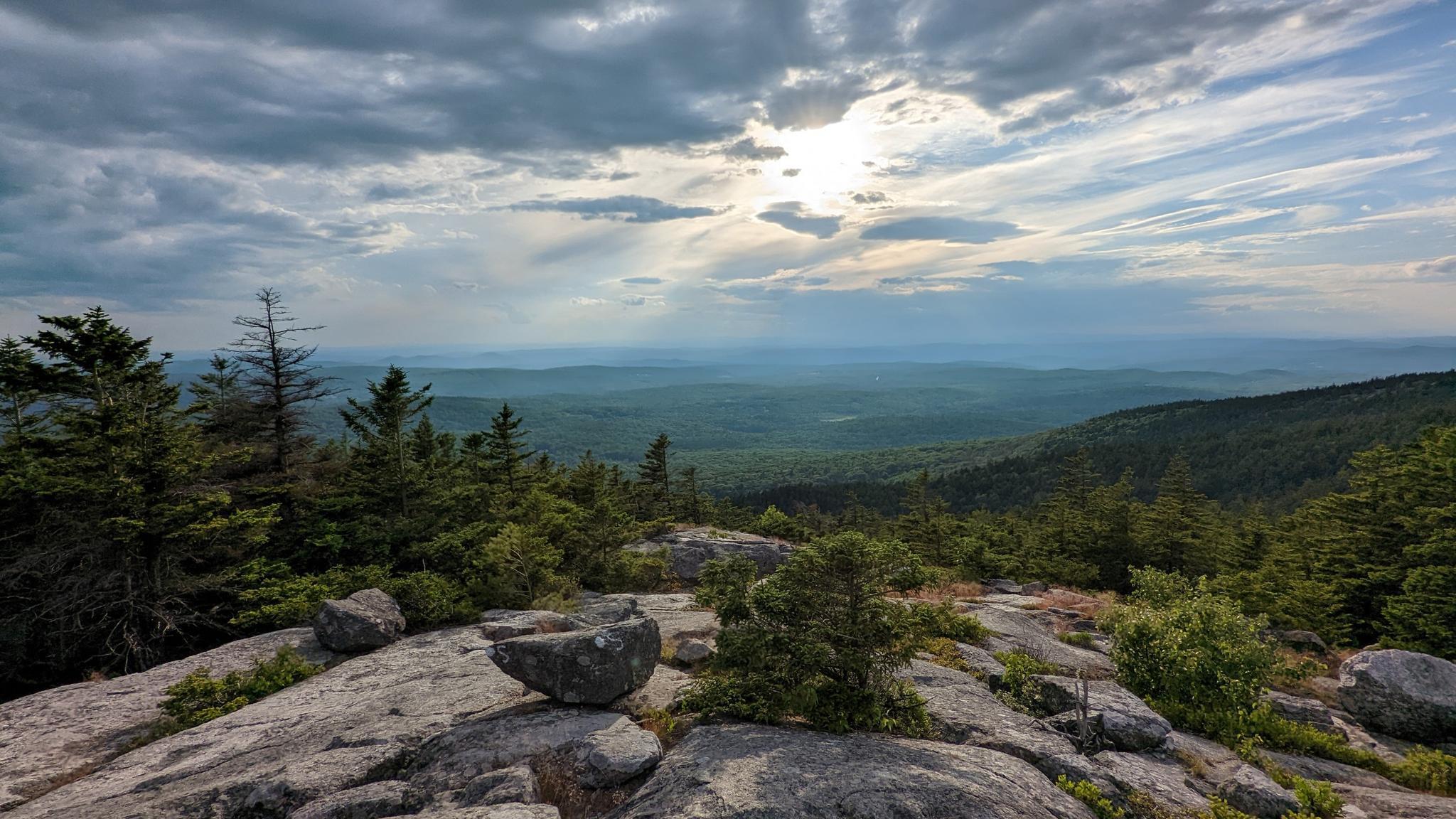 Expansive view from a rocky mountain top over dense green forests, cloudy sky.