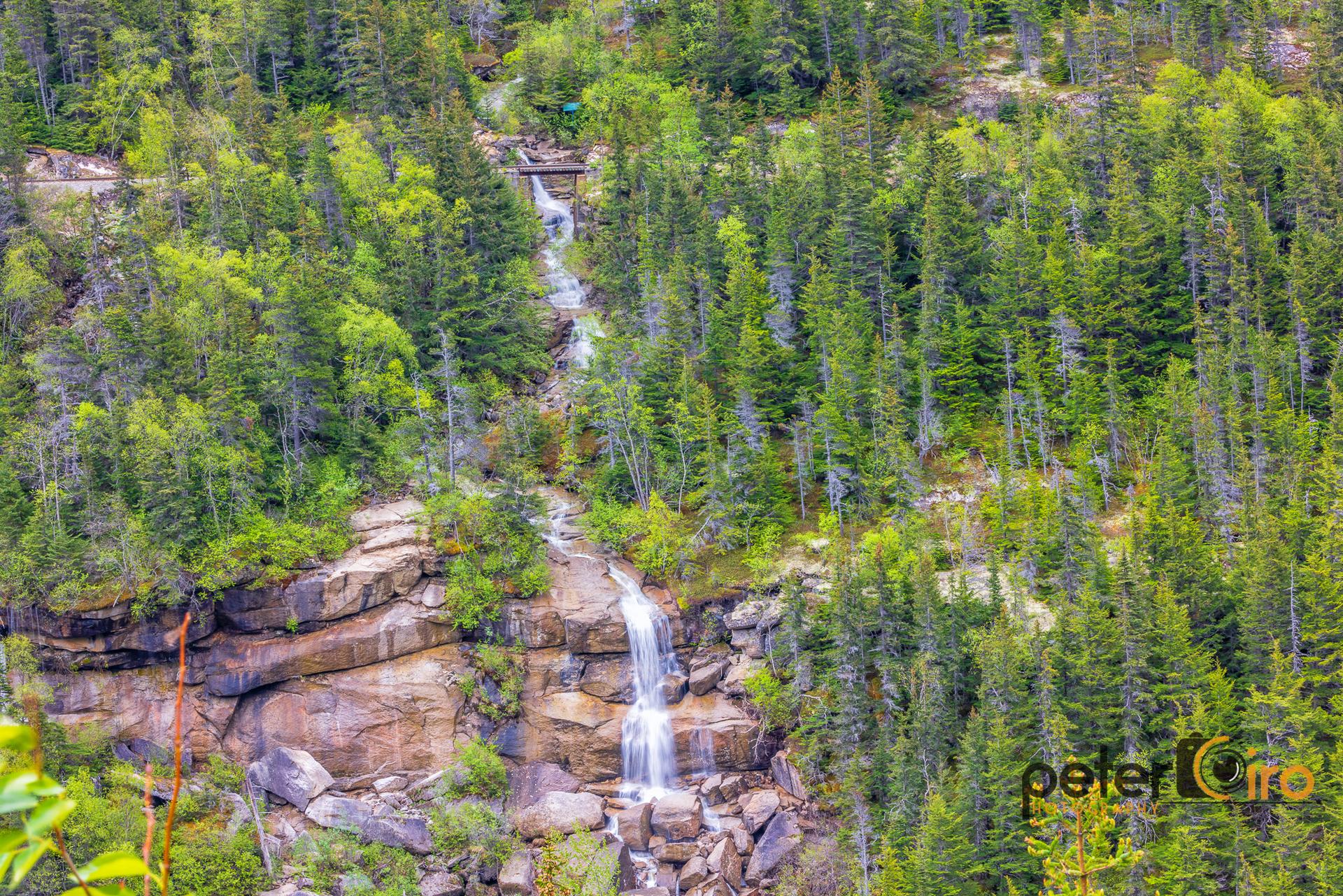 Multi-tiered waterfall cascades down rocky cliffs amidst a dense evergreen forest.