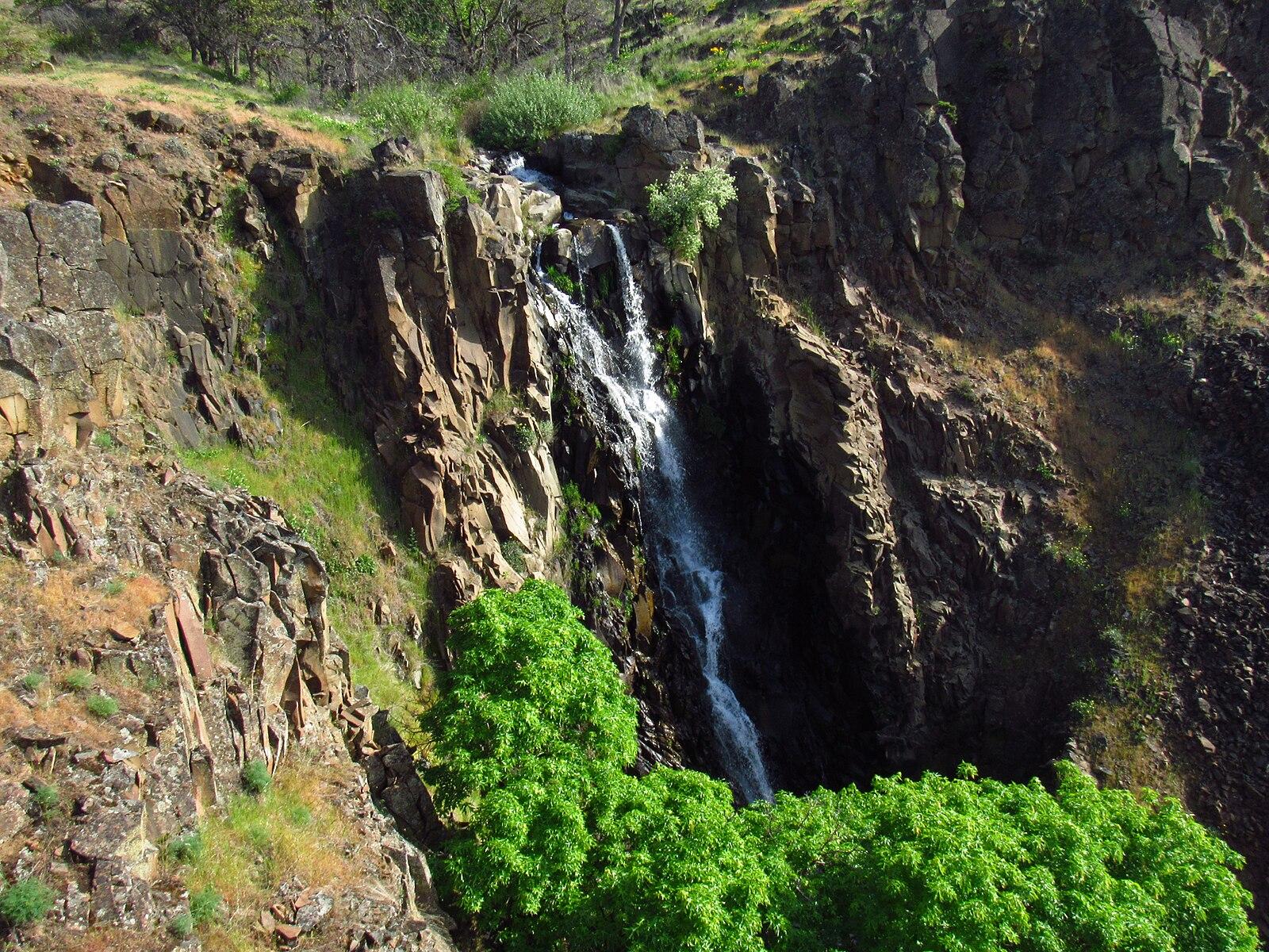 Waterfall cascades down a dark rocky cliff with bright green trees below.