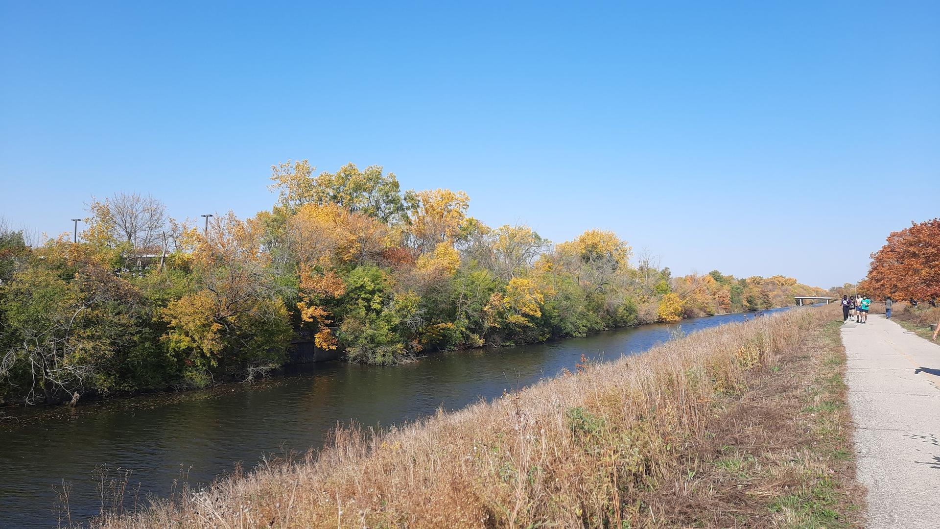 A sunny autumn day with a river, colorful trees, and people on a path.