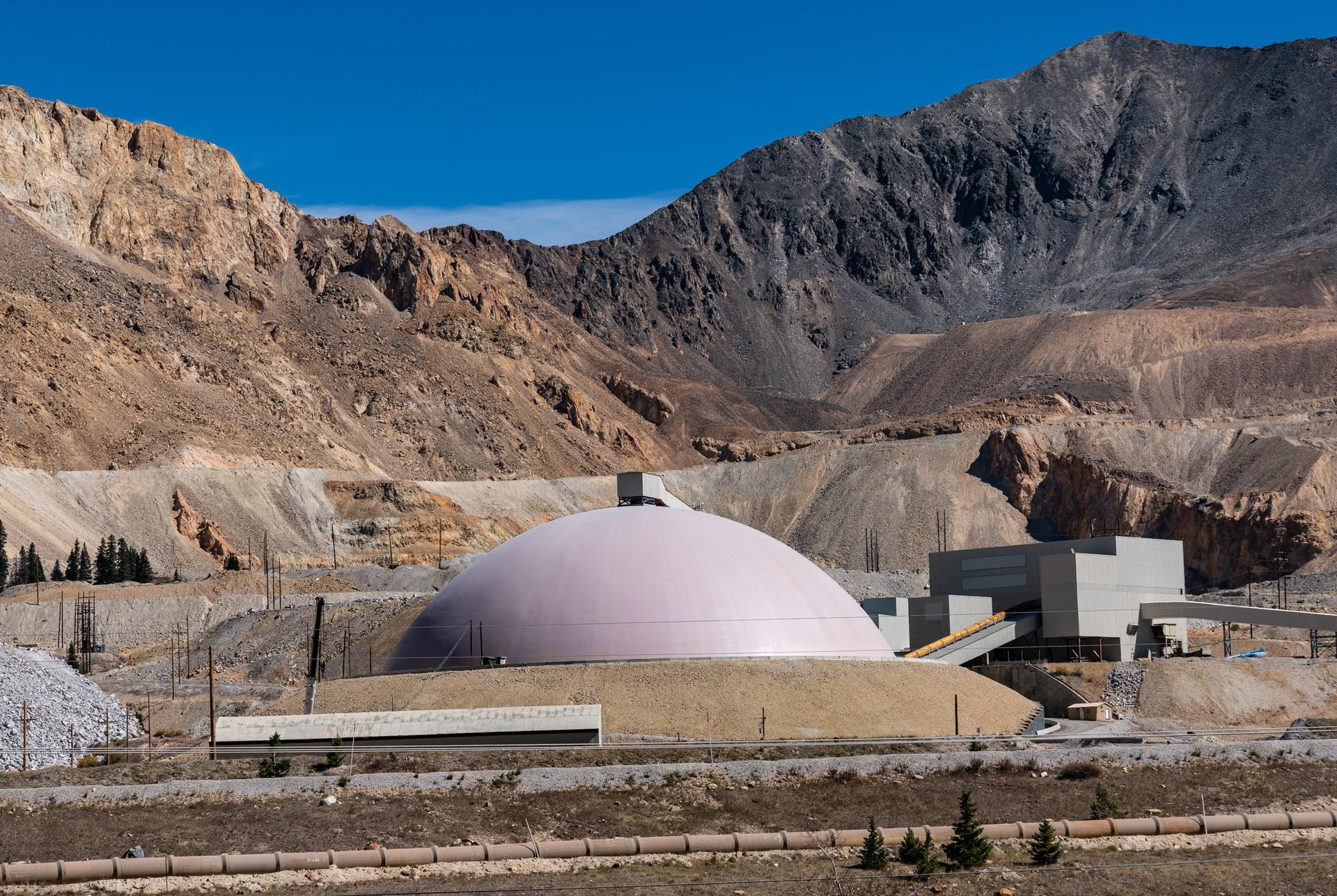 Large industrial dome and buildings against barren mountains under a blue sky.