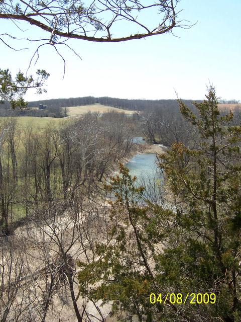 River winds through a valley with bare trees and evergreens under a bright sky.