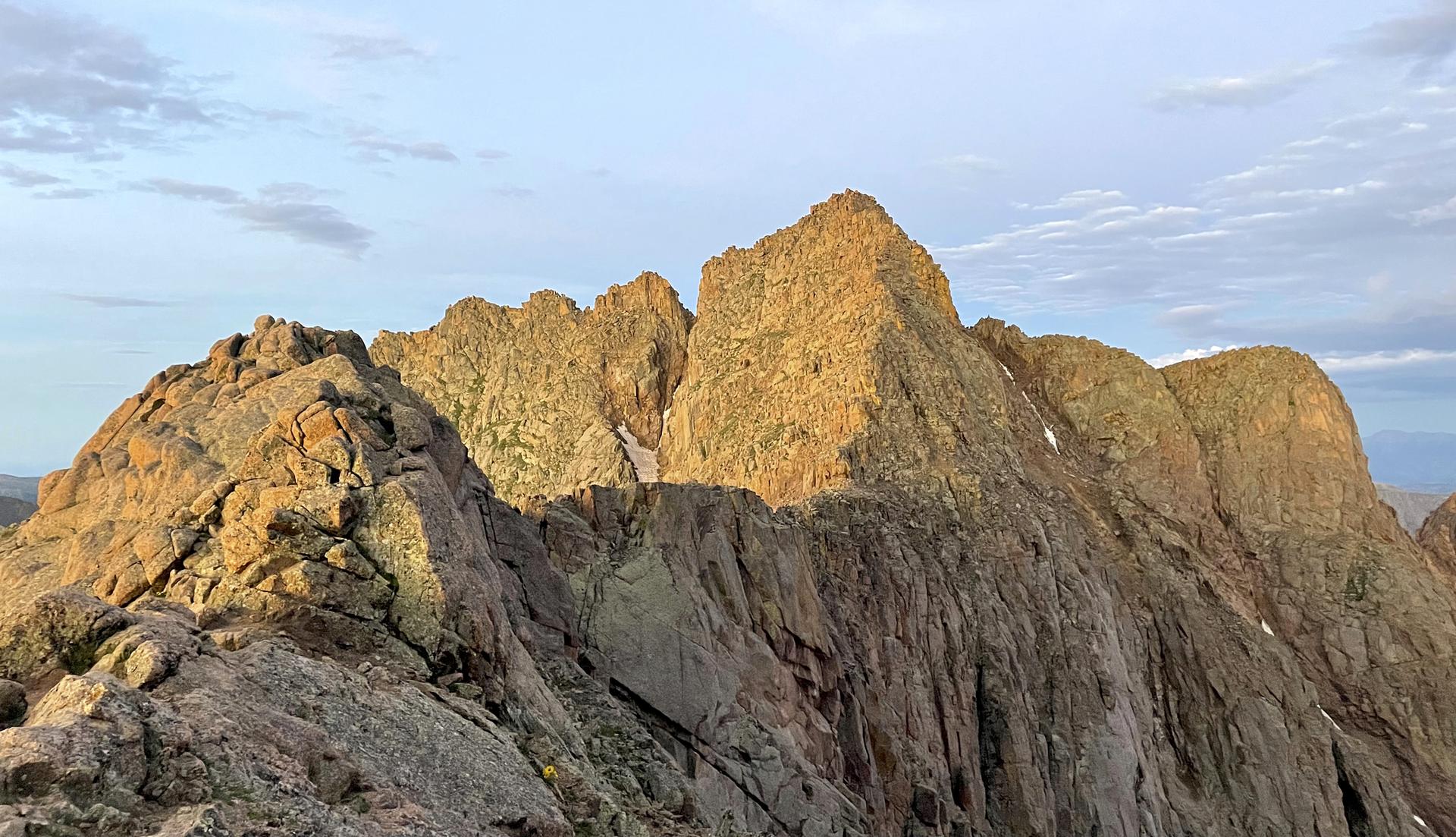 Golden light illuminates rugged mountain peaks with small snow patches under a soft sky.