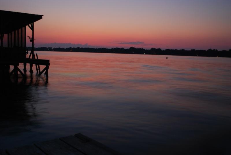 Silhouetted boathouse and dock at sunset with colorful sky reflecting on calm water.