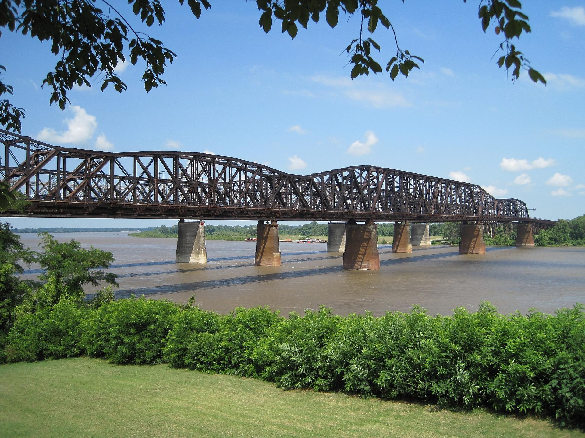 Large metal truss bridge spanning a wide river, framed by green trees.