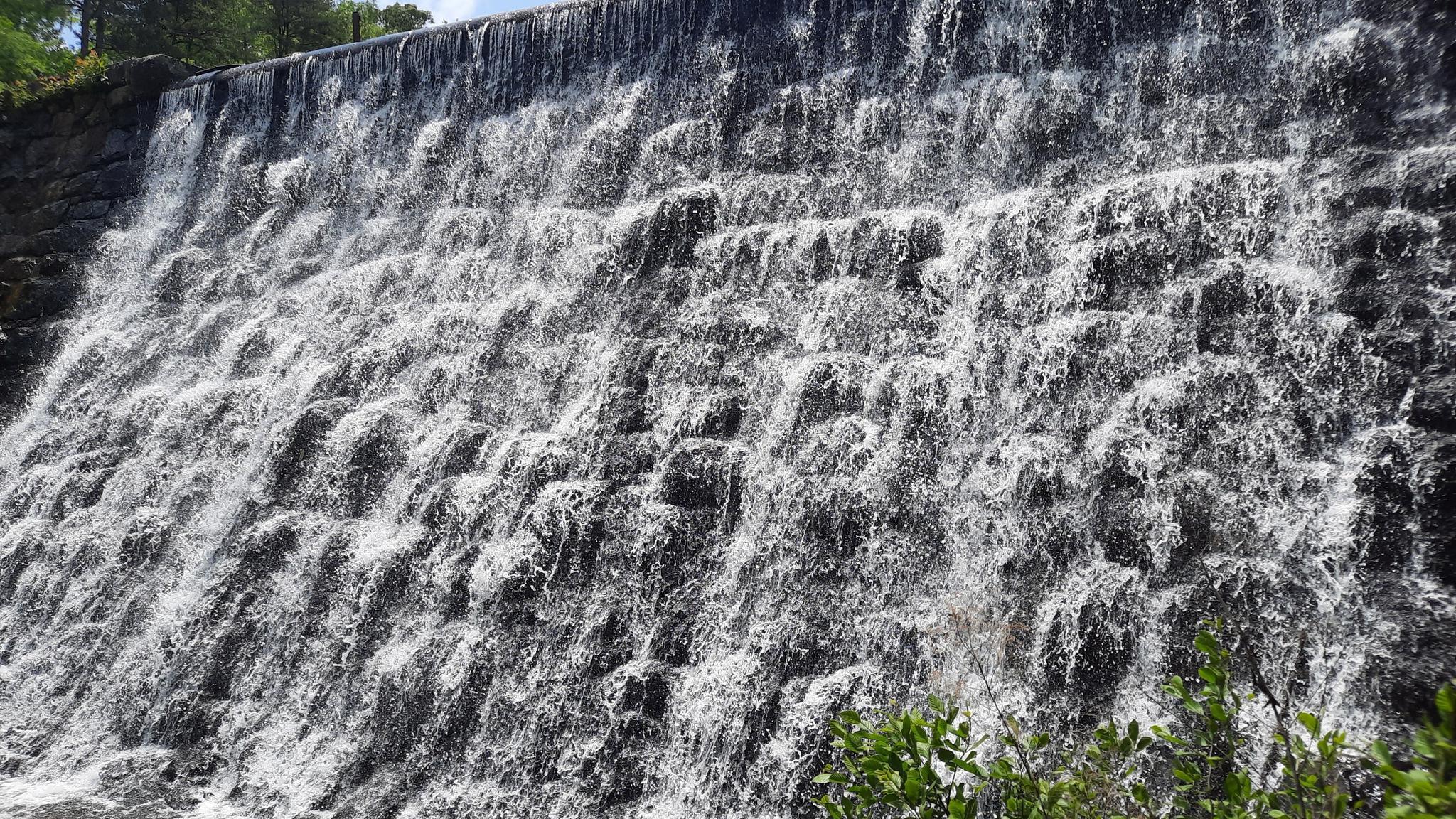 A wide waterfall cascades down a textured, man-made wall.