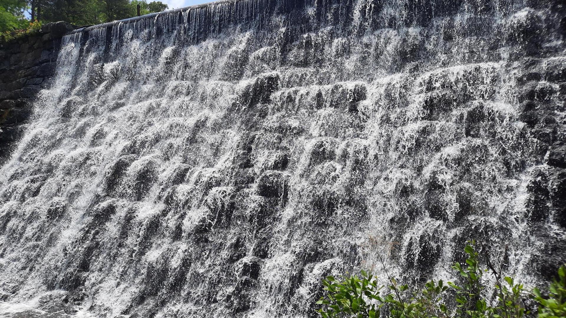 A wide waterfall cascades down a textured, man-made wall.