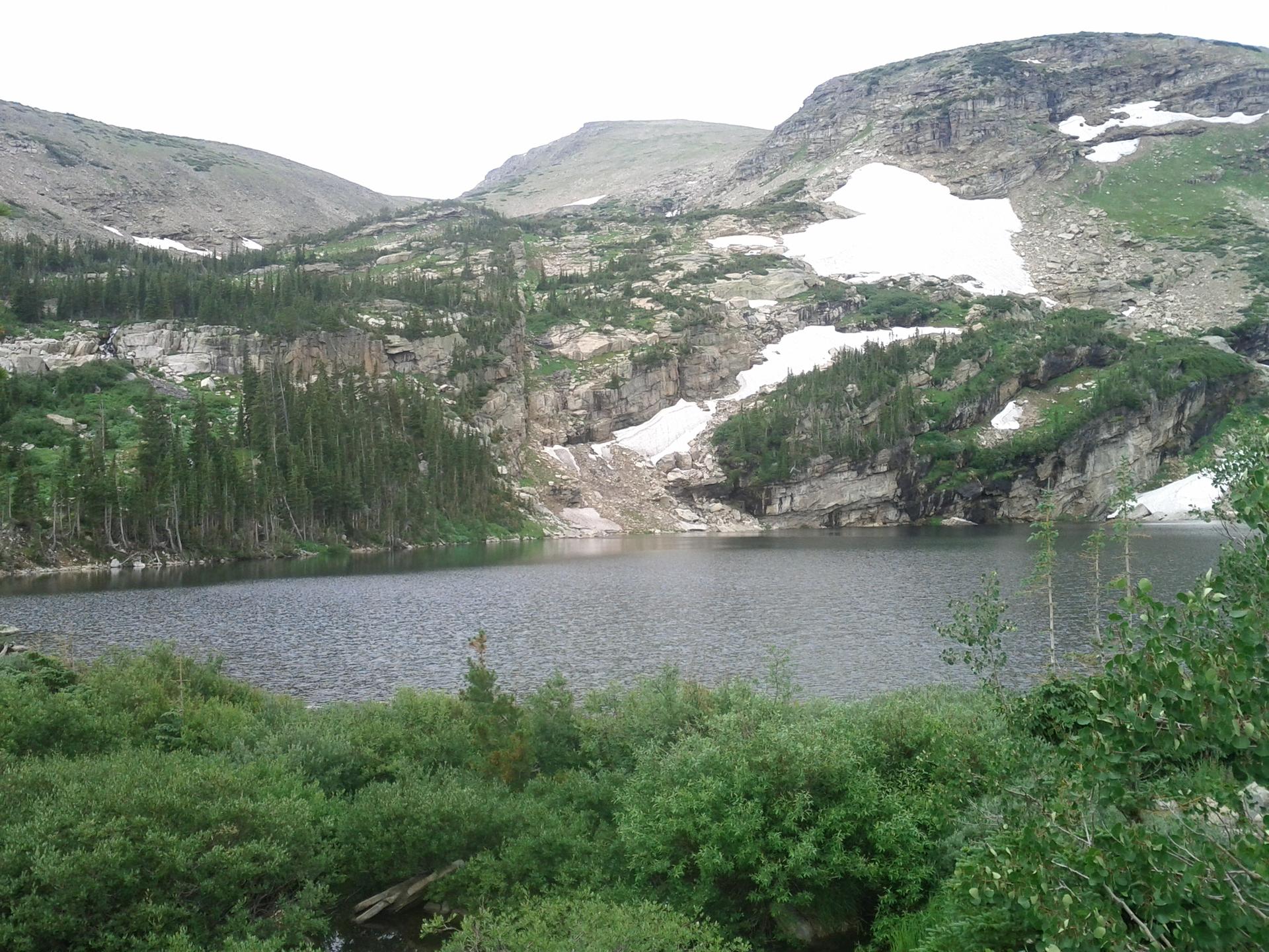 A serene mountain lake surrounded by green trees and snow under an overcast sky.