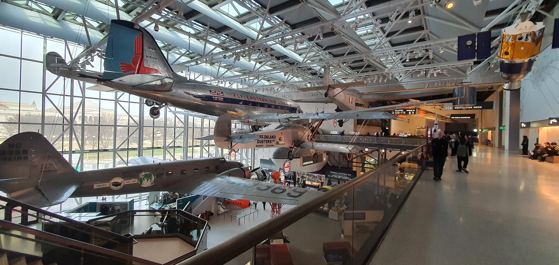 Indoor museum with suspended airplanes, people walking below.