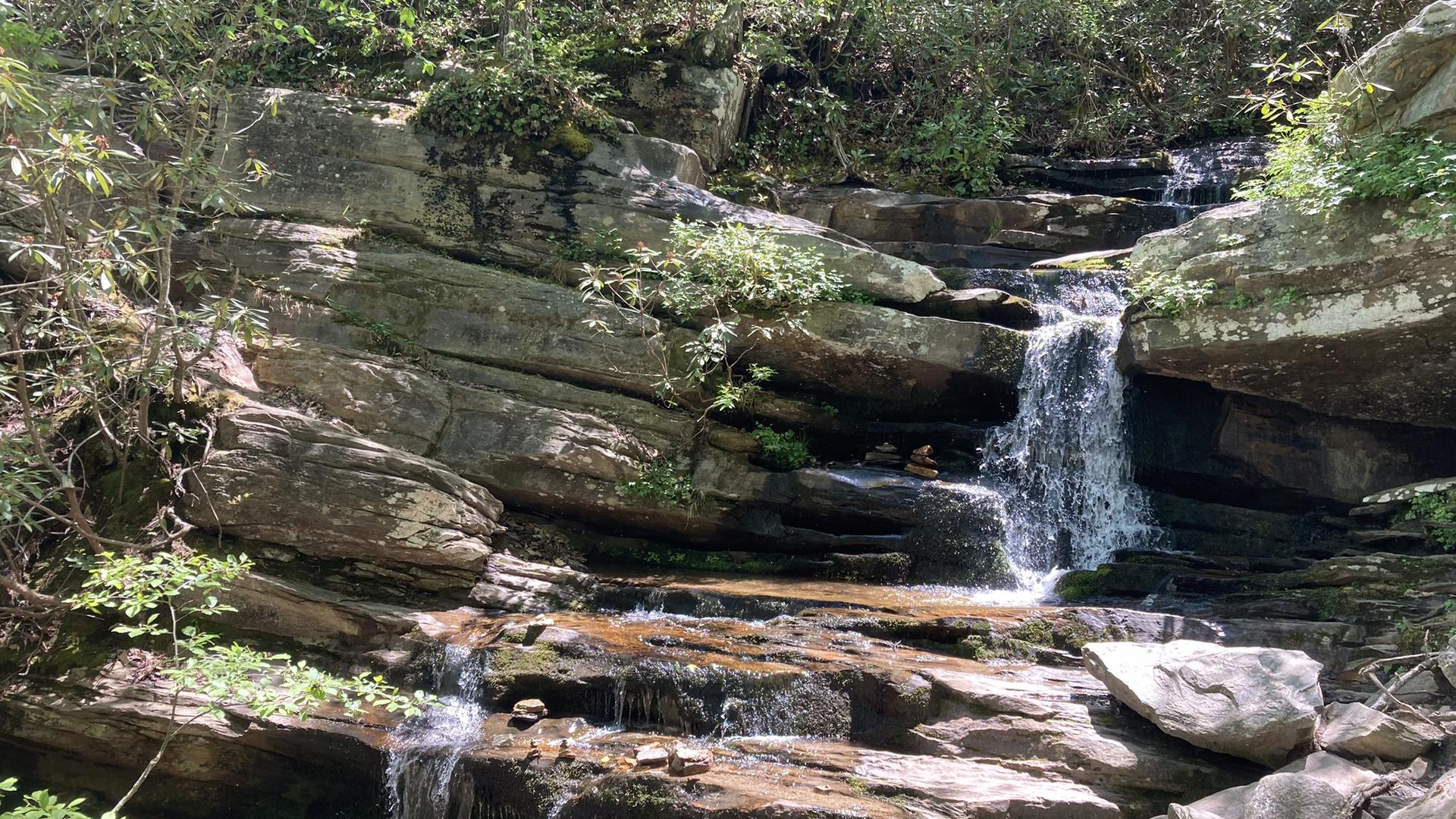 A multi-tiered waterfall flows over sunlit rocks amidst lush green trees.