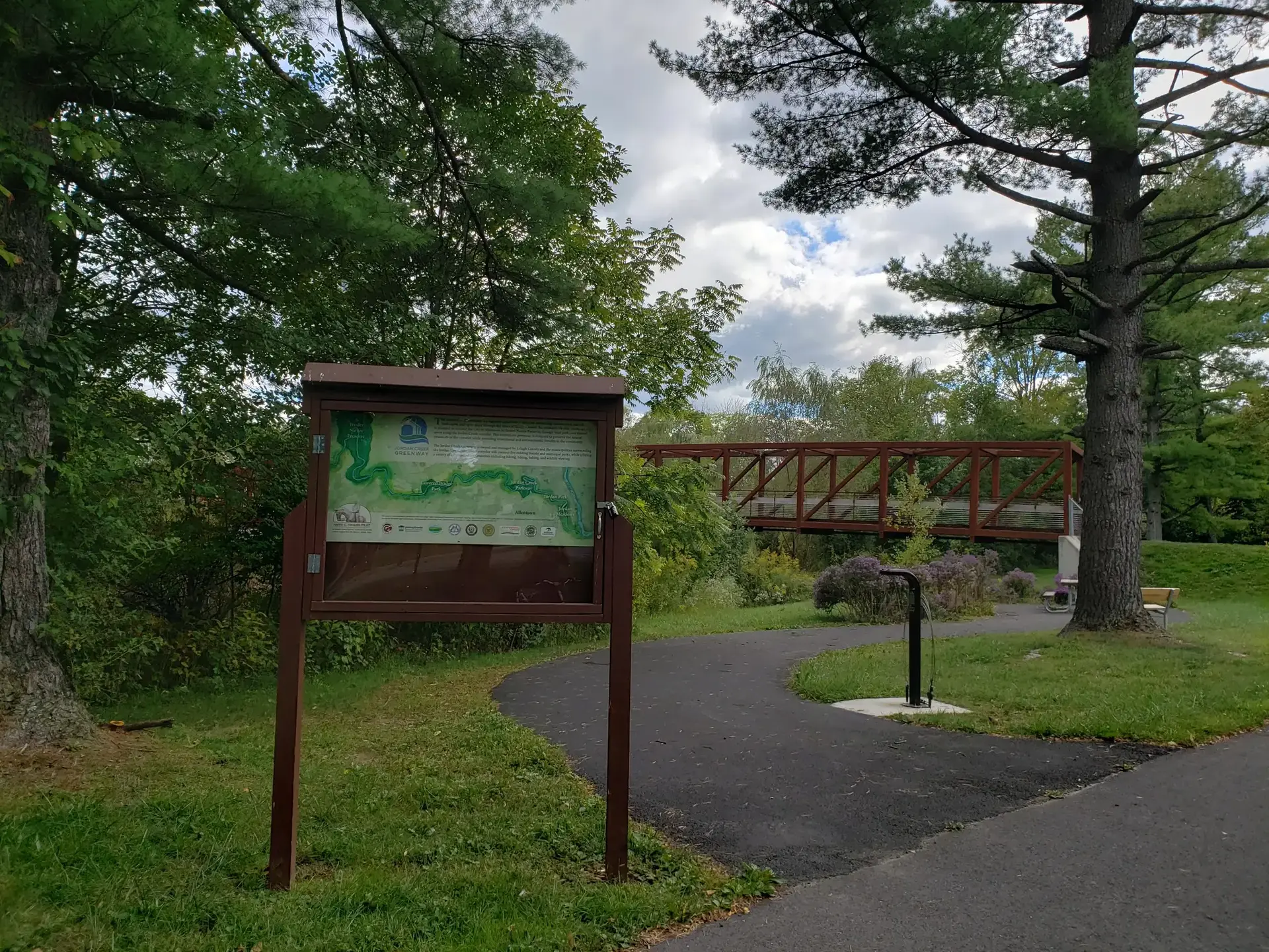 A trail map stands beside a paved path leading to a metal bridge.