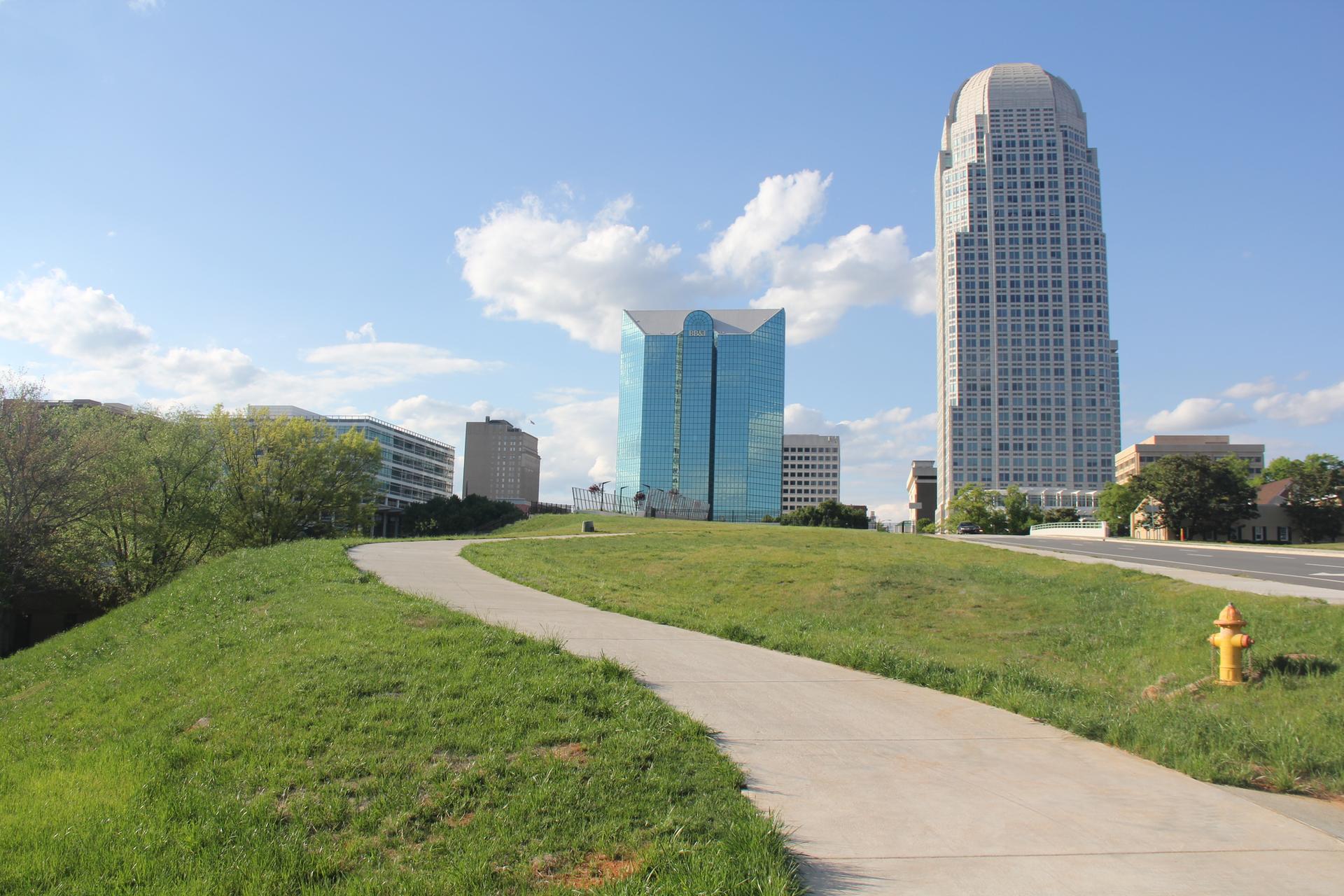 A paved path winds up a grassy hill towards city buildings under blue sky.