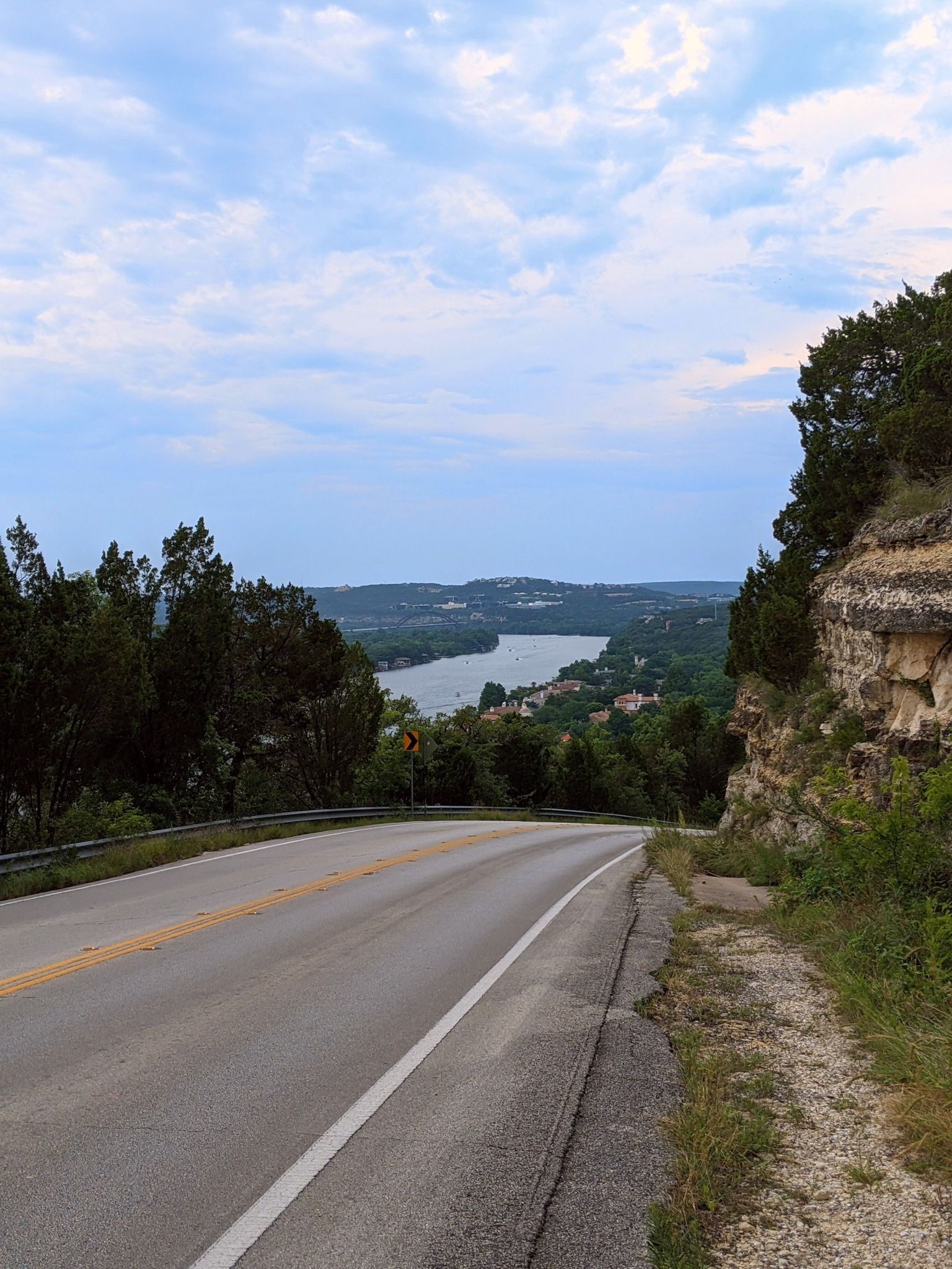 Curving road next to a rocky cliff overlooking a wide river and hills. - Mount Bonnell Trail