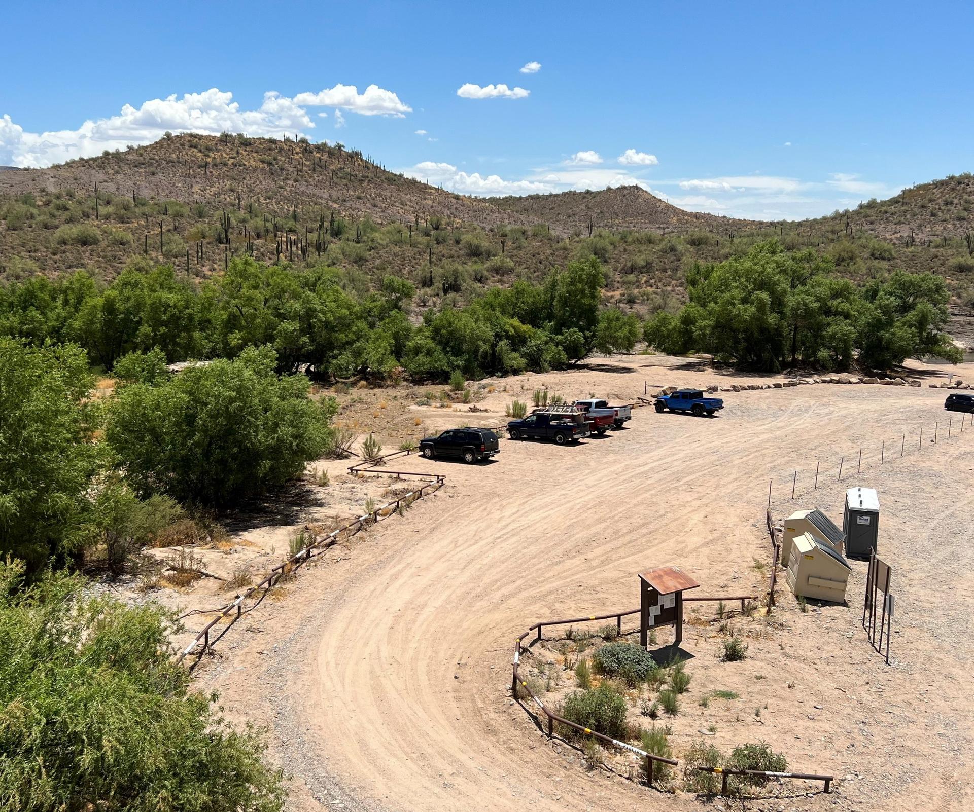Dirt parking lot with trucks and trailers in a sunny, arid, hilly landscape.