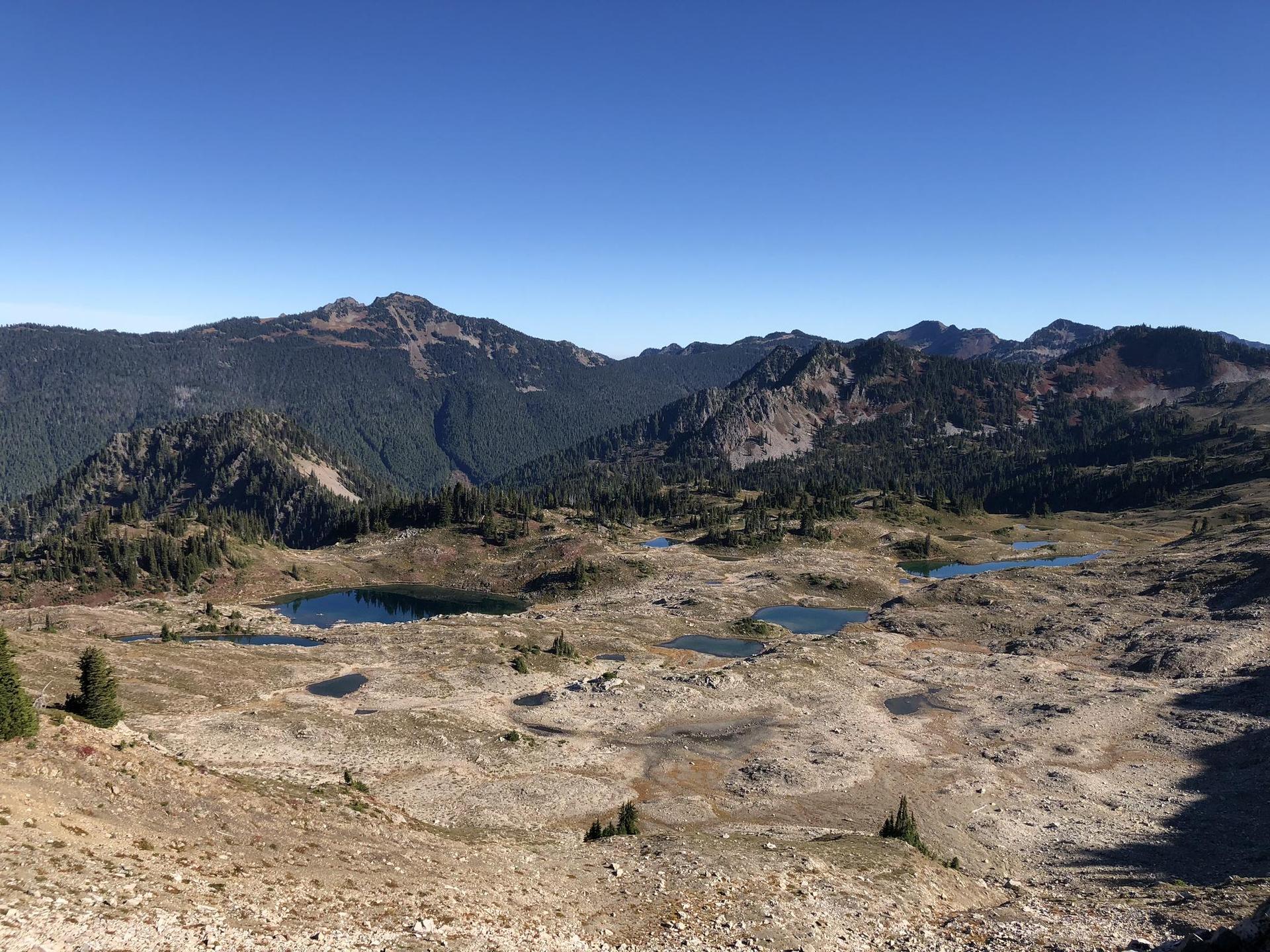 Expansive view of rocky alpine terrain with small lakes and distant forested mountains.