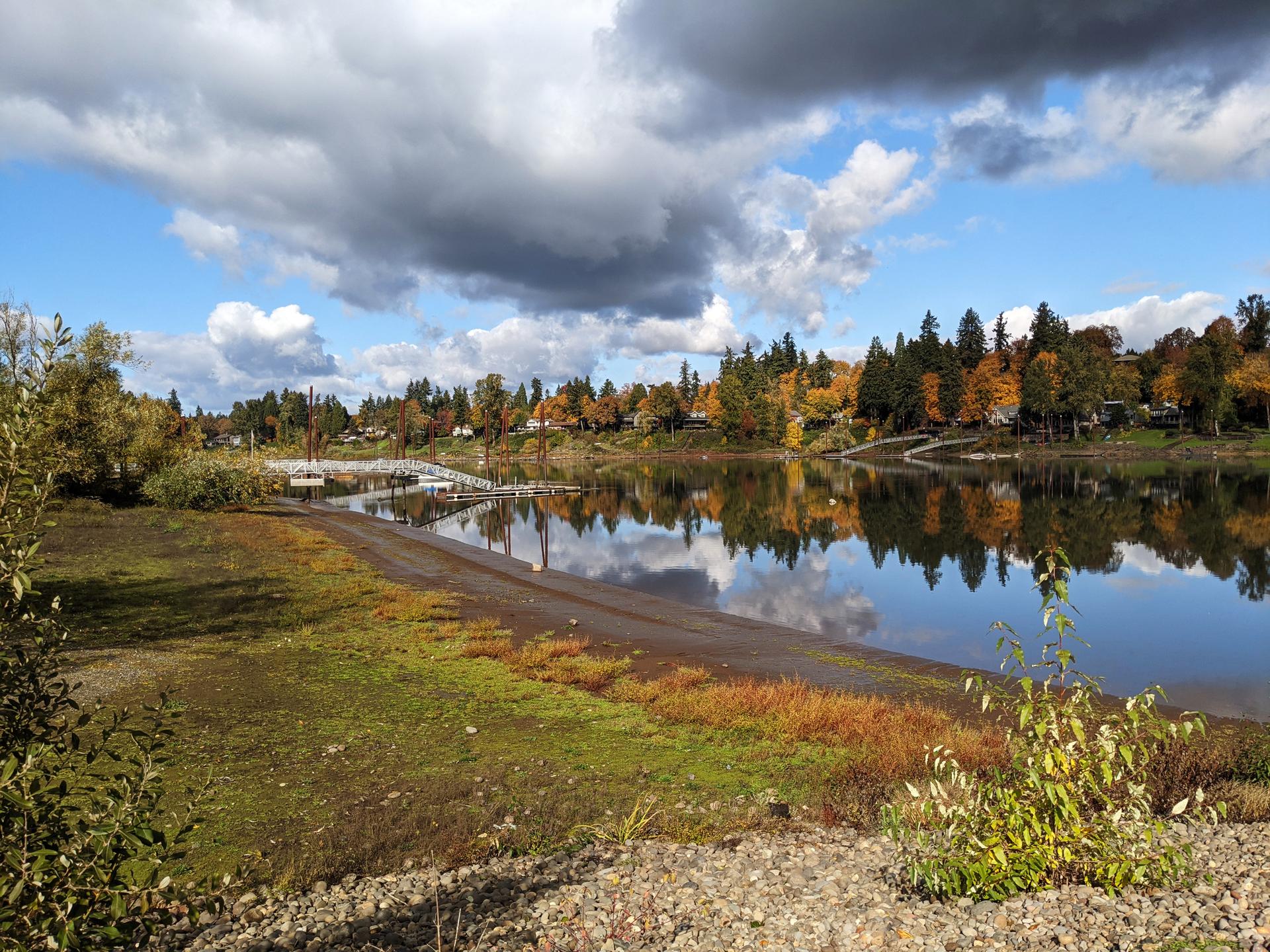 Calm river reflecting autumn trees and cloudy sky, with distant docks.