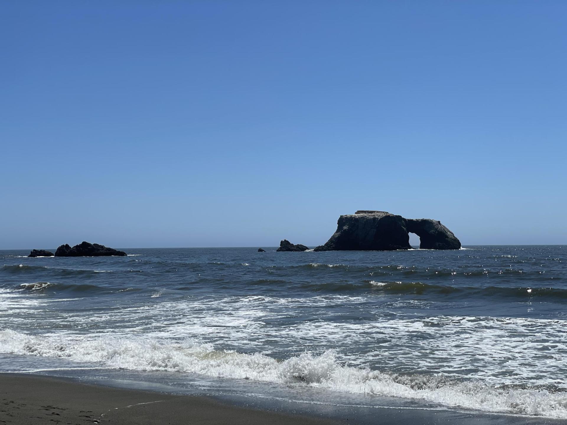 Bright ocean with a large sea arch rock and waves on a sandy beach.