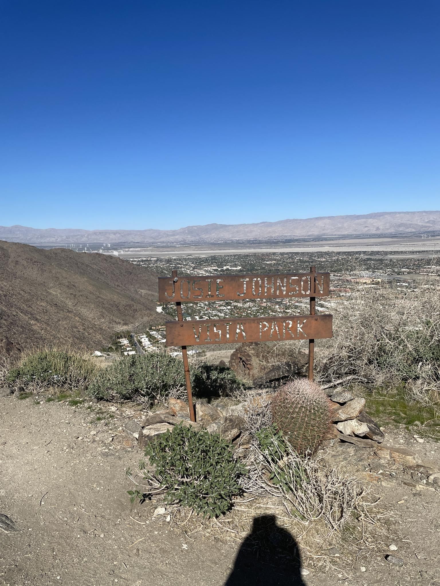 Rusty sign on a desert hillside, overlooking a valley town, with a person's shadow. - South Carl Lykken Trail - North Section