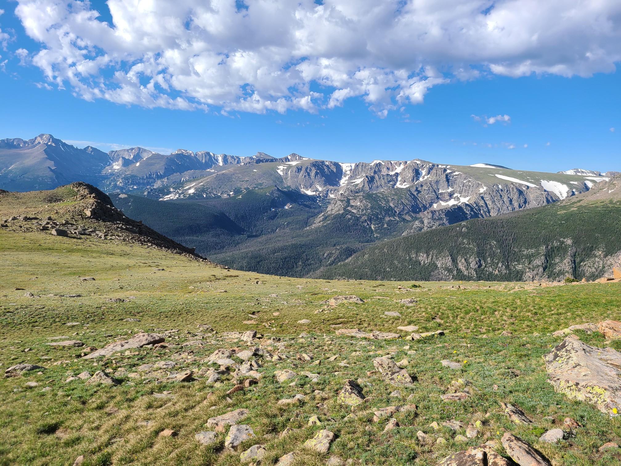 Green, rocky hills overlook snow-capped mountains under a bright blue sky.