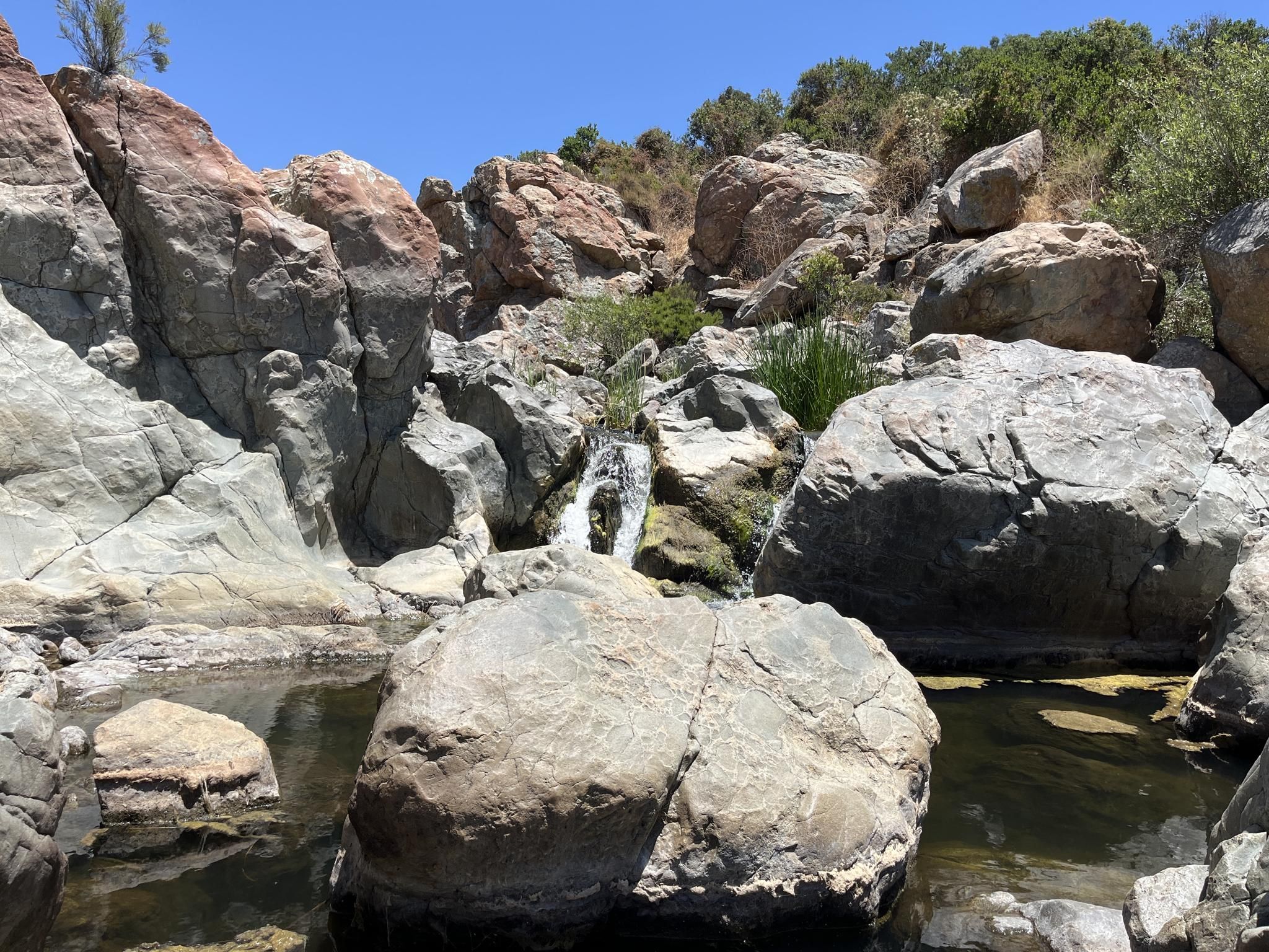 A small waterfall cascades into a clear pool surrounded by large rocks. - Los Peñasquitos Falls loop from Lopez Canyon