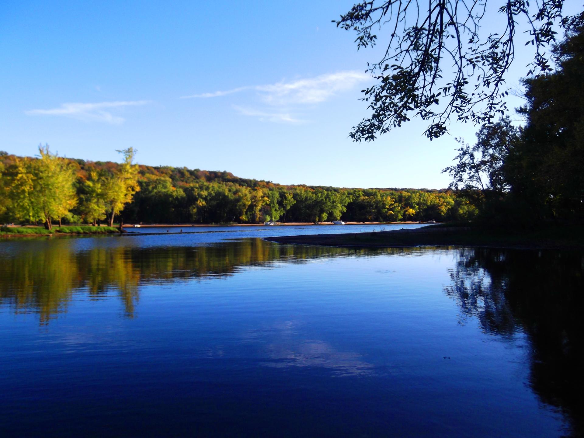 Wide, calm river reflecting green and yellow trees under a clear blue sky.