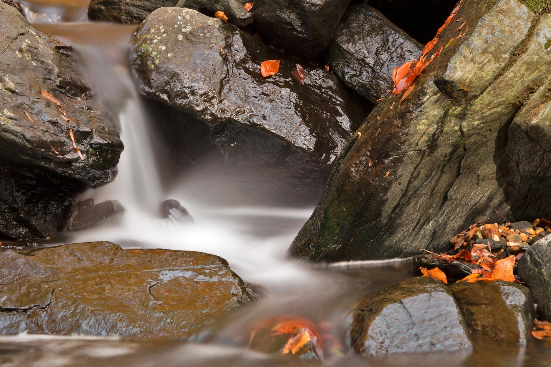 Silky waterfall cascades over dark rocks with scattered orange autumn leaves.