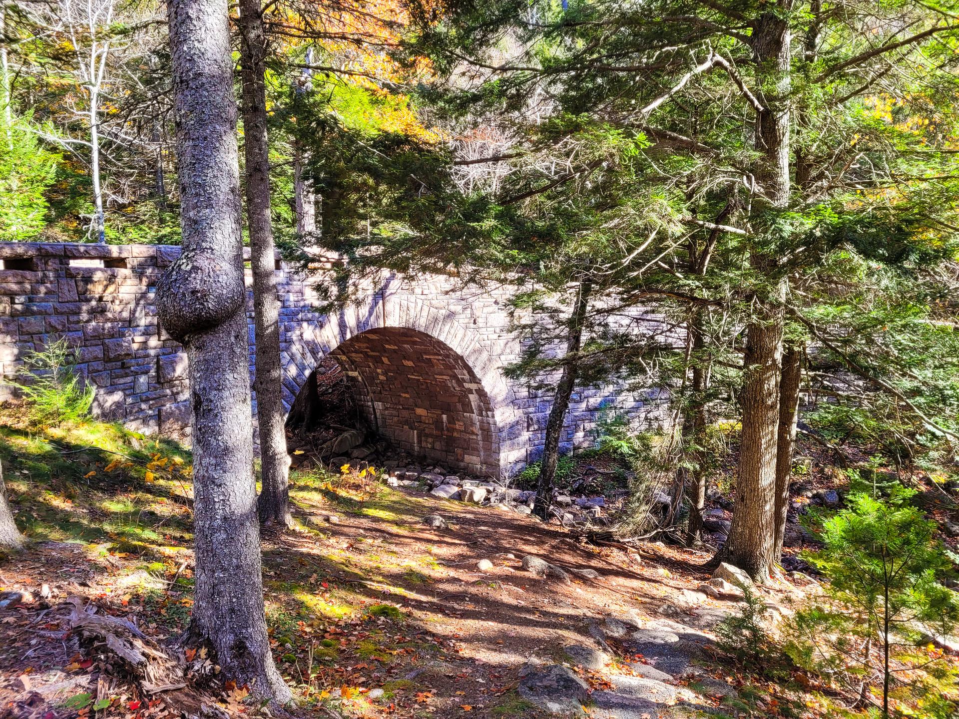 Stone arch bridge in a sun-dappled forest with autumn colors.