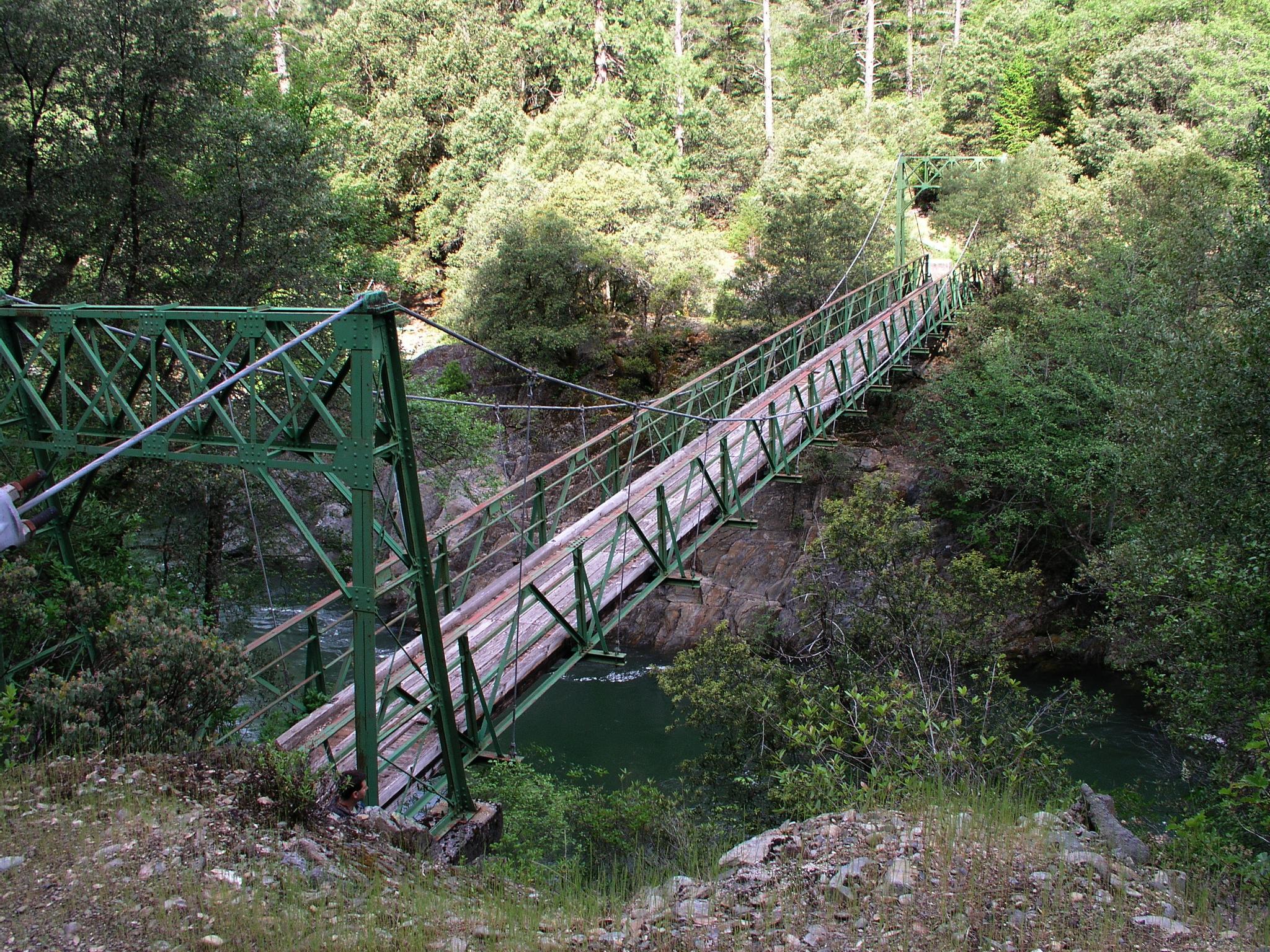 Long green suspension bridge spans a river through a dense forest.