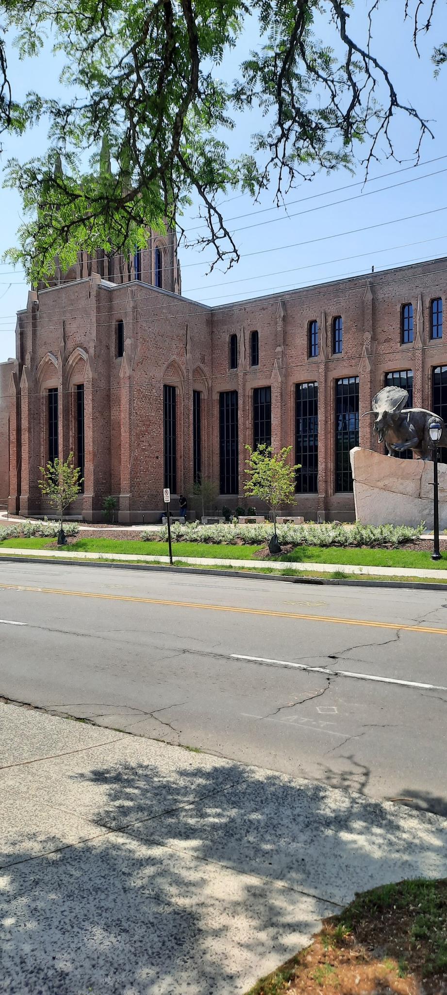 Large brick building with dome, street, and green landscaping on a sunny day.